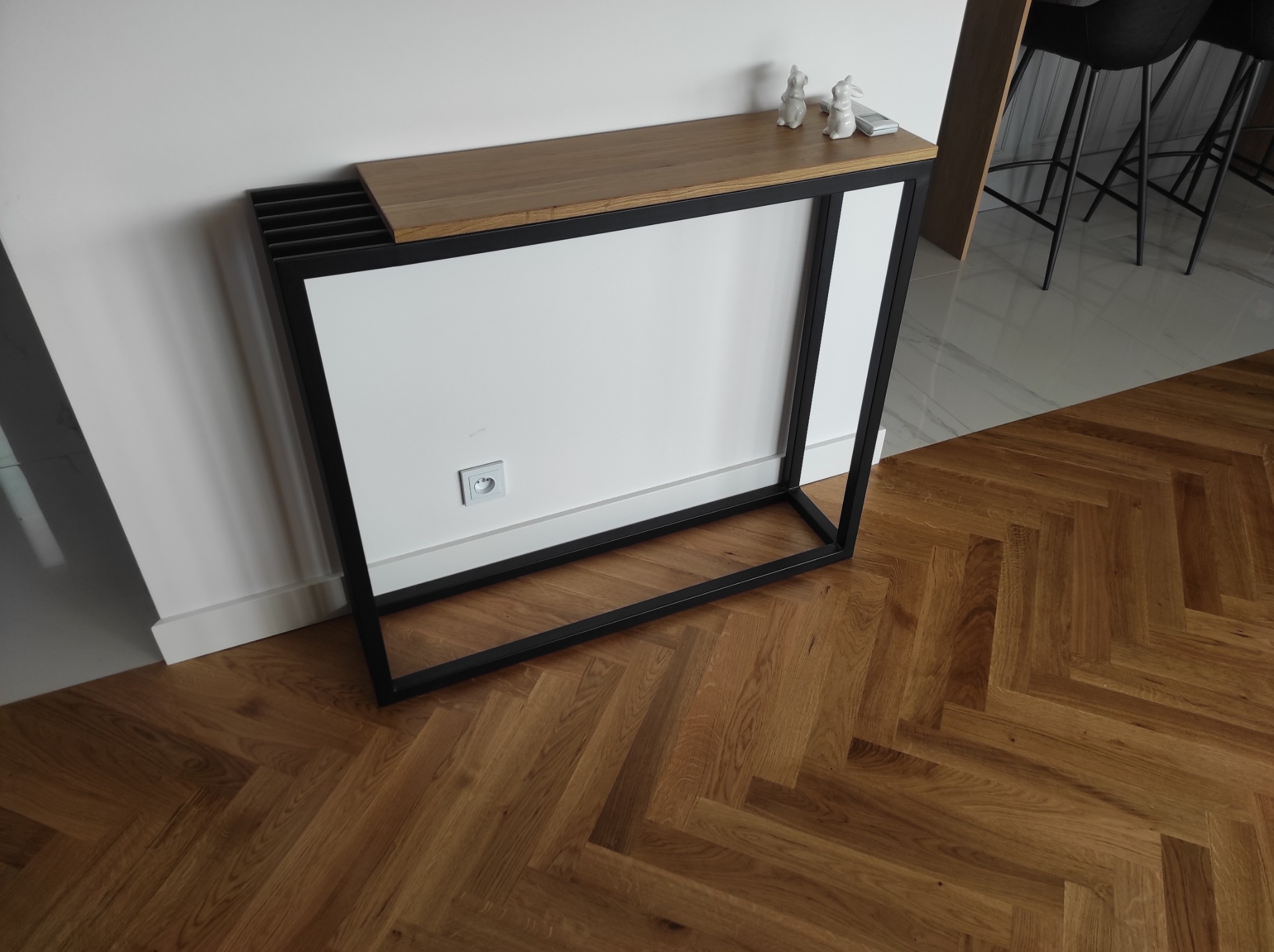 Metalowa konsola z drewnianym blatem, białą osłoną grzejnika i dekoracjami w kształcie królików, ustawiona na parkiecie w jodełkę.