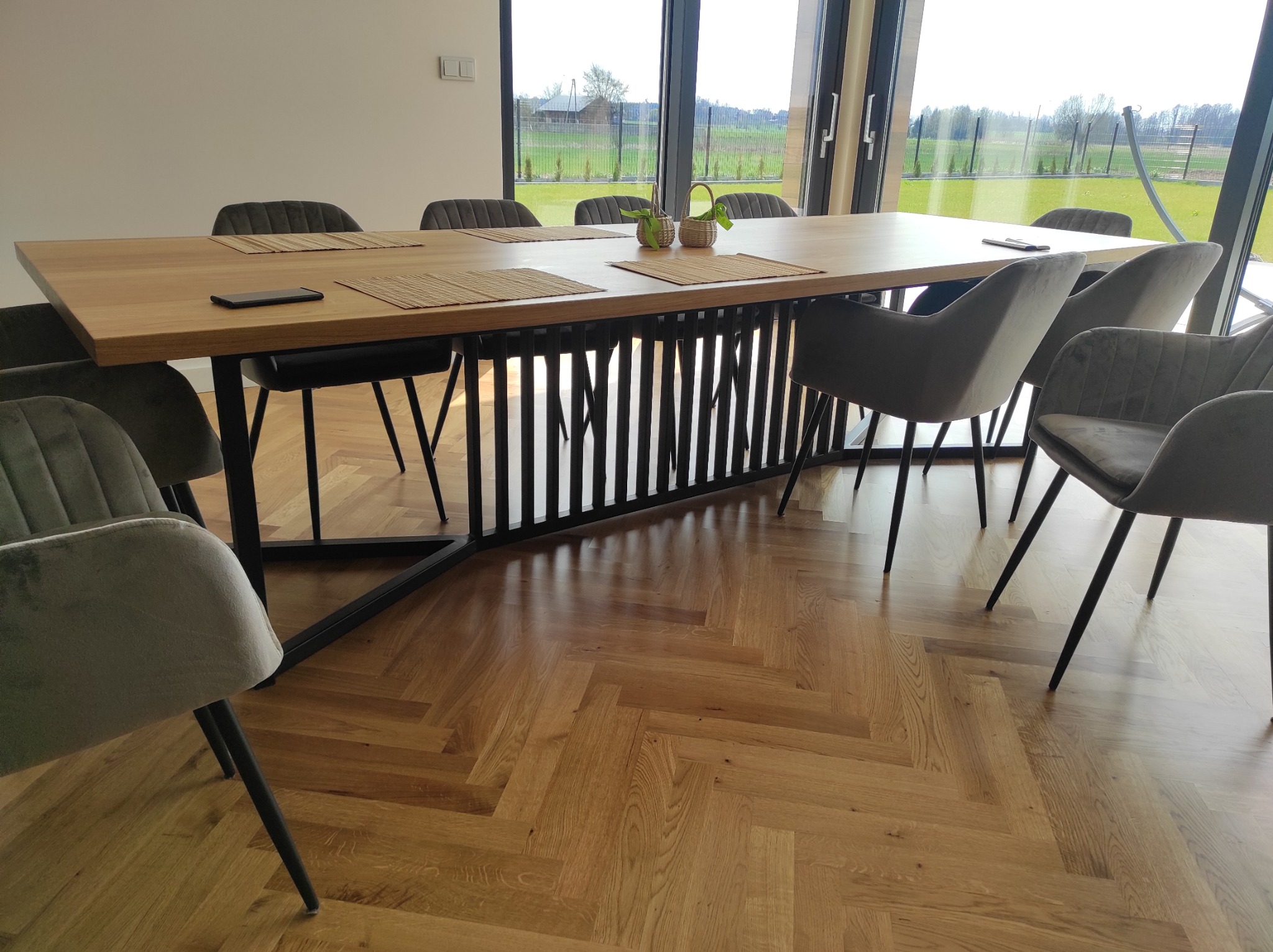 Stół z drewnianym blatem i metalową podstawą z pionowych prętów, otoczony szarymi krzesłami na tle dużego okna z widokiem na zielony krajobraz.