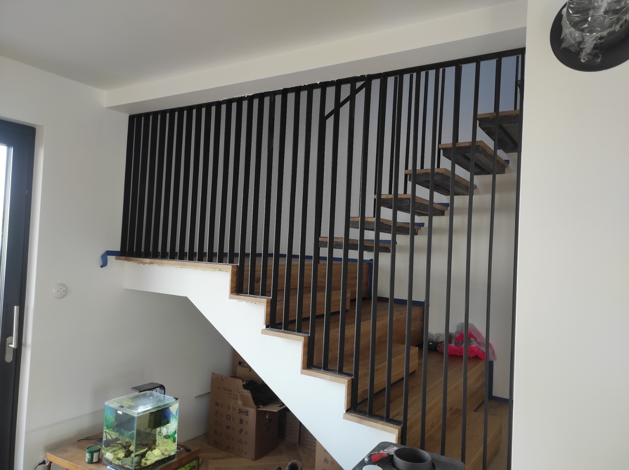 Wnętrze domu w trakcie wykańczania: schody z drewnianymi stopniami i czarną, pionową balustradą, widoczne kartony, akwarium i zabezpieczenia malarskie.