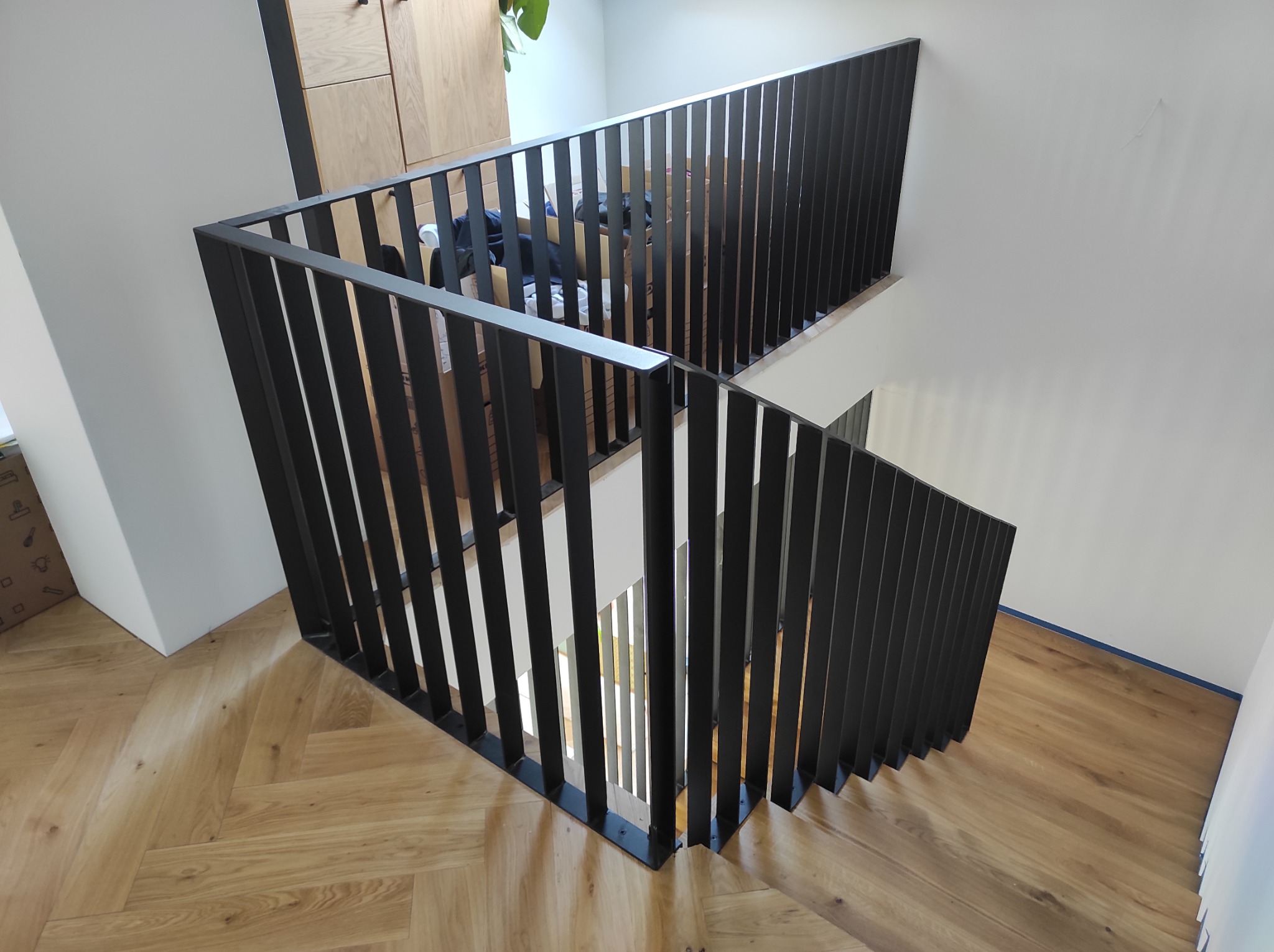 Nowoczesna, czarna balustrada schodowa z pionowych, płaskich prętów, zamontowana na drewnianych schodach ułożonych w jodełkę, widok z góry.
