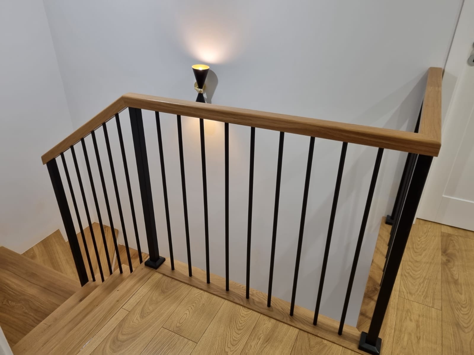 Nowoczesna balustrada schodowa z drewnianym pochwytem i czarnymi, pionowymi prętami, oświetlona lampą ścienną w kształcie stożka, widok z góry na drewniane schody.