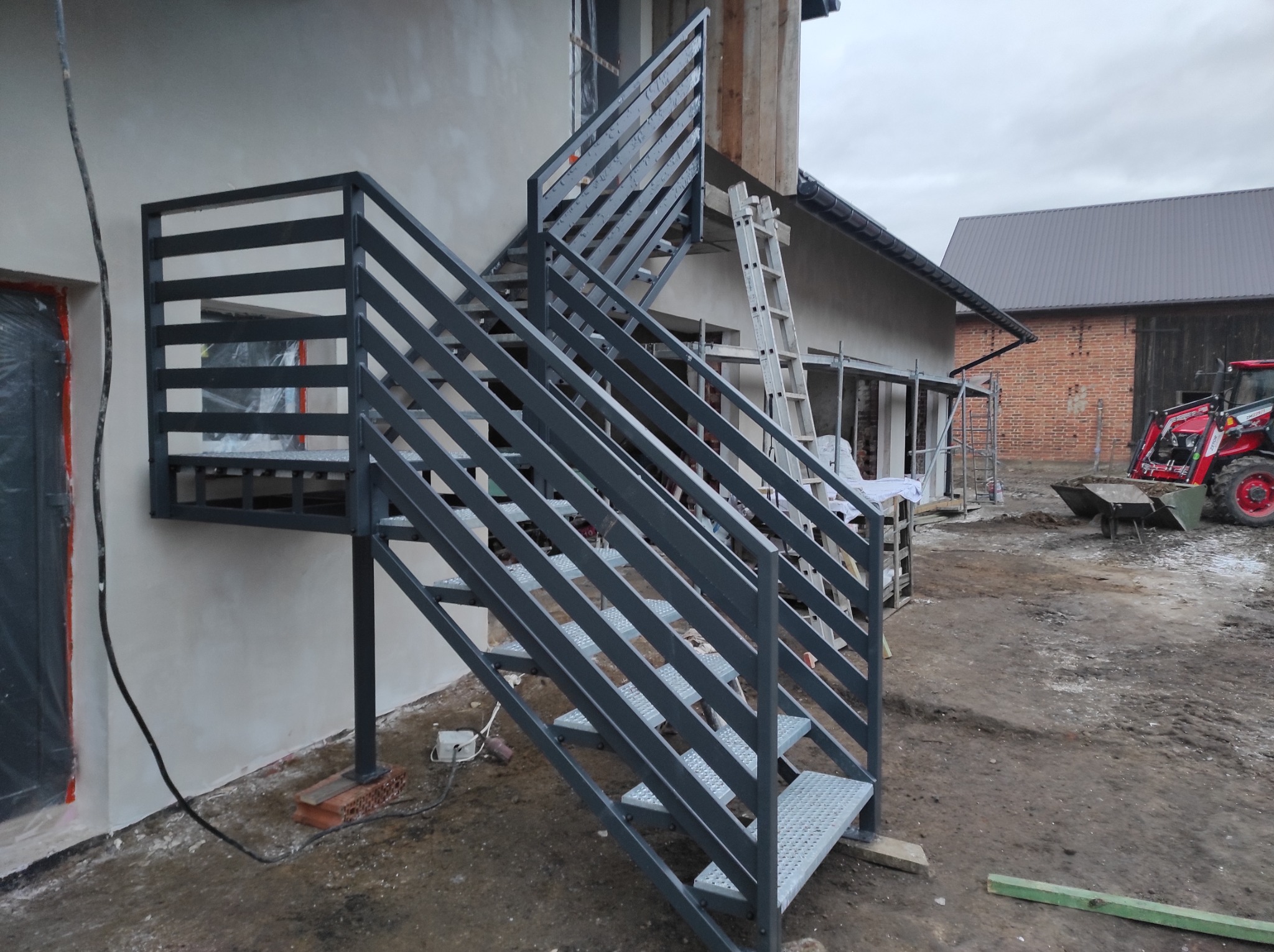 Zewnętrzne schody metalowe z szarymi poręczami prowadzące do wejścia budynku w trakcie budowy, widoczny traktor i rusztowania.