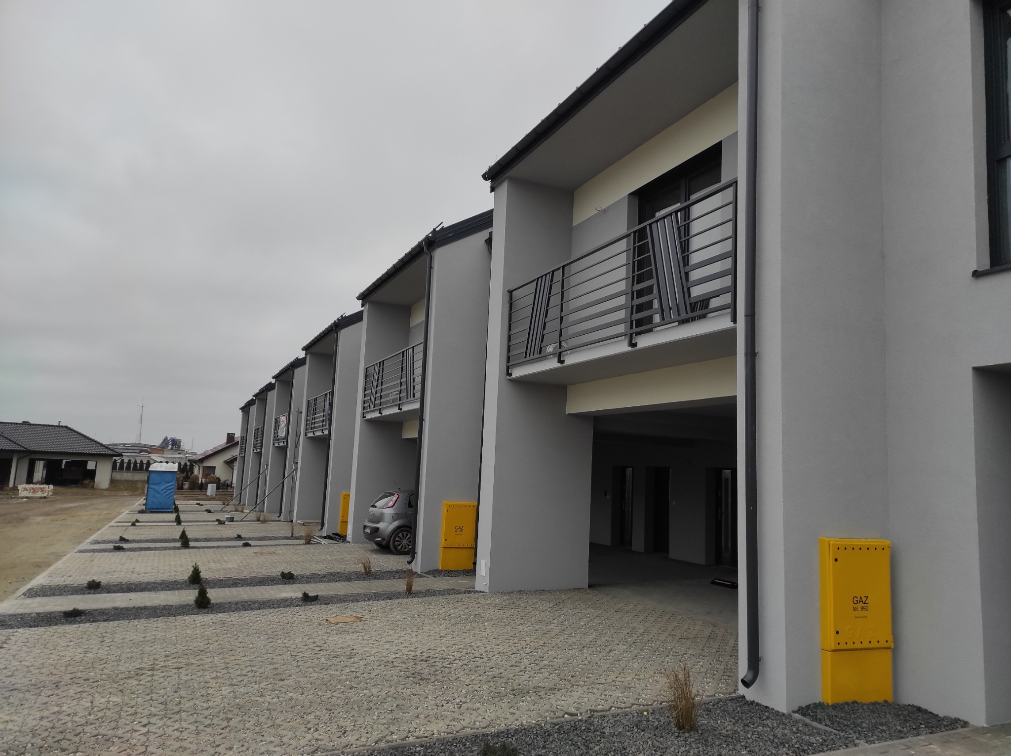 Szereg nowych, szarych budynków mieszkalnych z balkonami z czarnymi, poziomymi barierkami. Na pierwszym planie wyłożony kostką brukową podjazd z małymi drzewkami i żółtymi skrzynkami gazowymi.