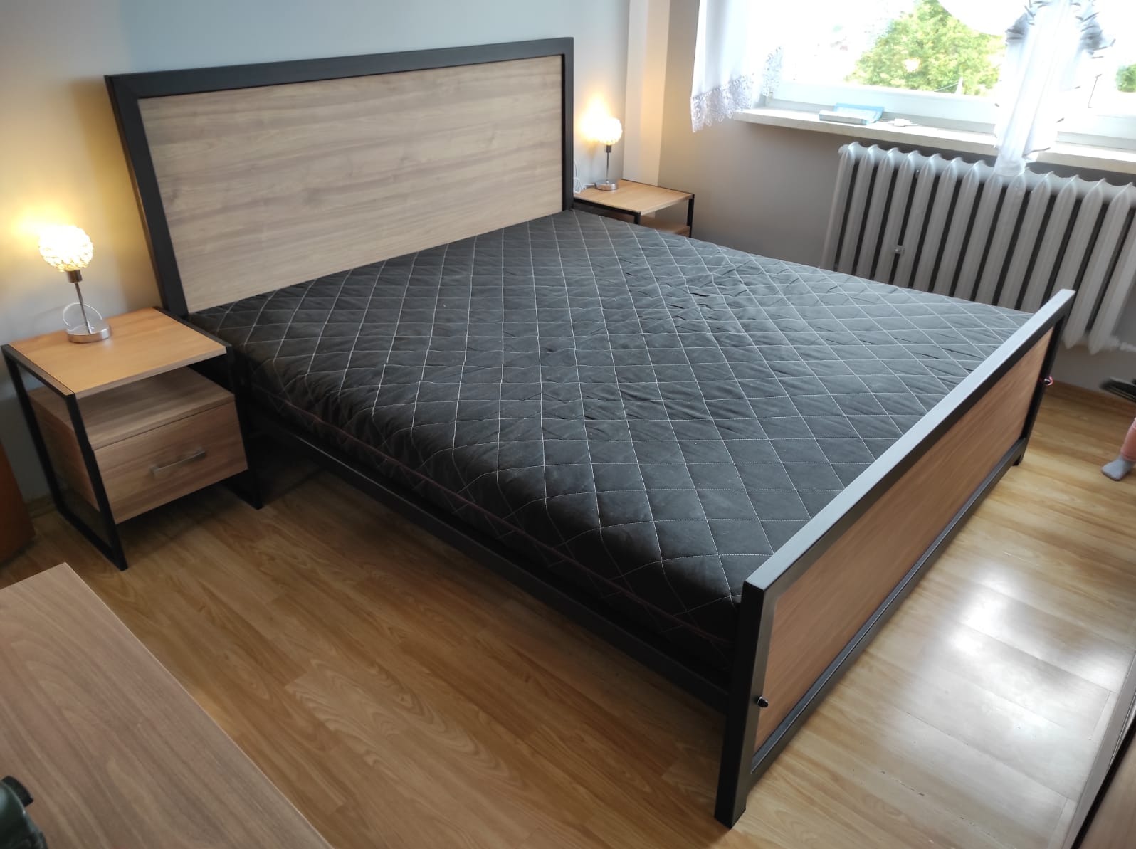 Nowoczesne łóżko z metalową ramą i drewnianym wezgłowiem, ustawione w sypialni z drewnianą podłogą i dwoma stolikami nocnymi z lampkami.