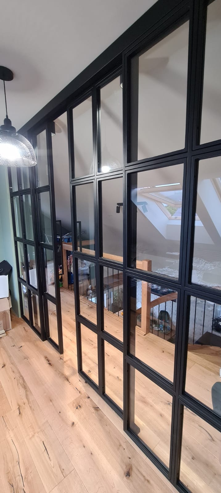 Przeszklona ścianka działowa z czarnymi metalowymi profilami, oddzielająca przestrzeń na dwóch poziomach, widok na schody i okno dachowe.