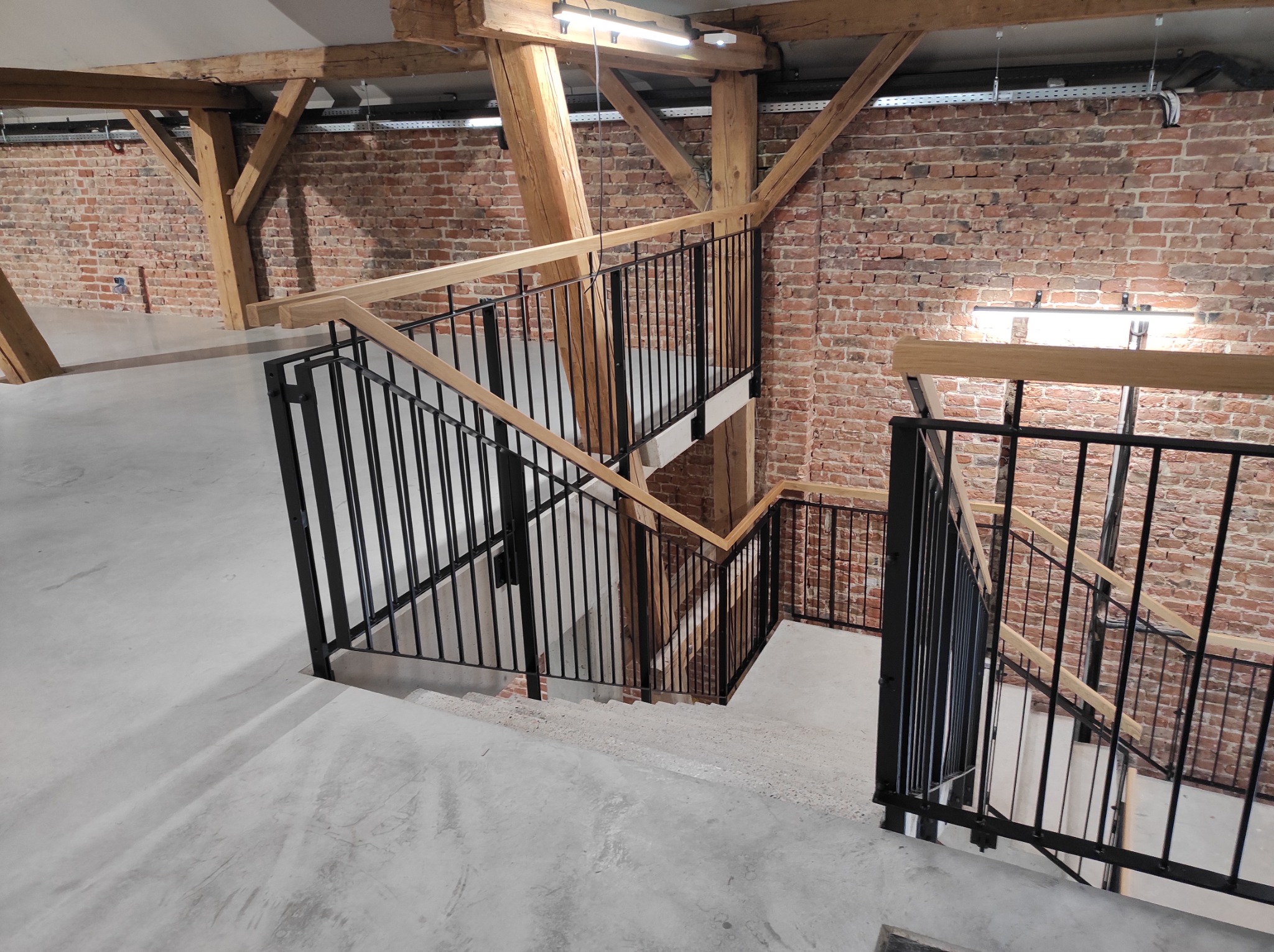 Wnętrze z betonowymi schodami i czarnymi metalowymi balustradami z drewnianymi poręczami, widoczne drewniane belki stropowe i ceglana ściana.