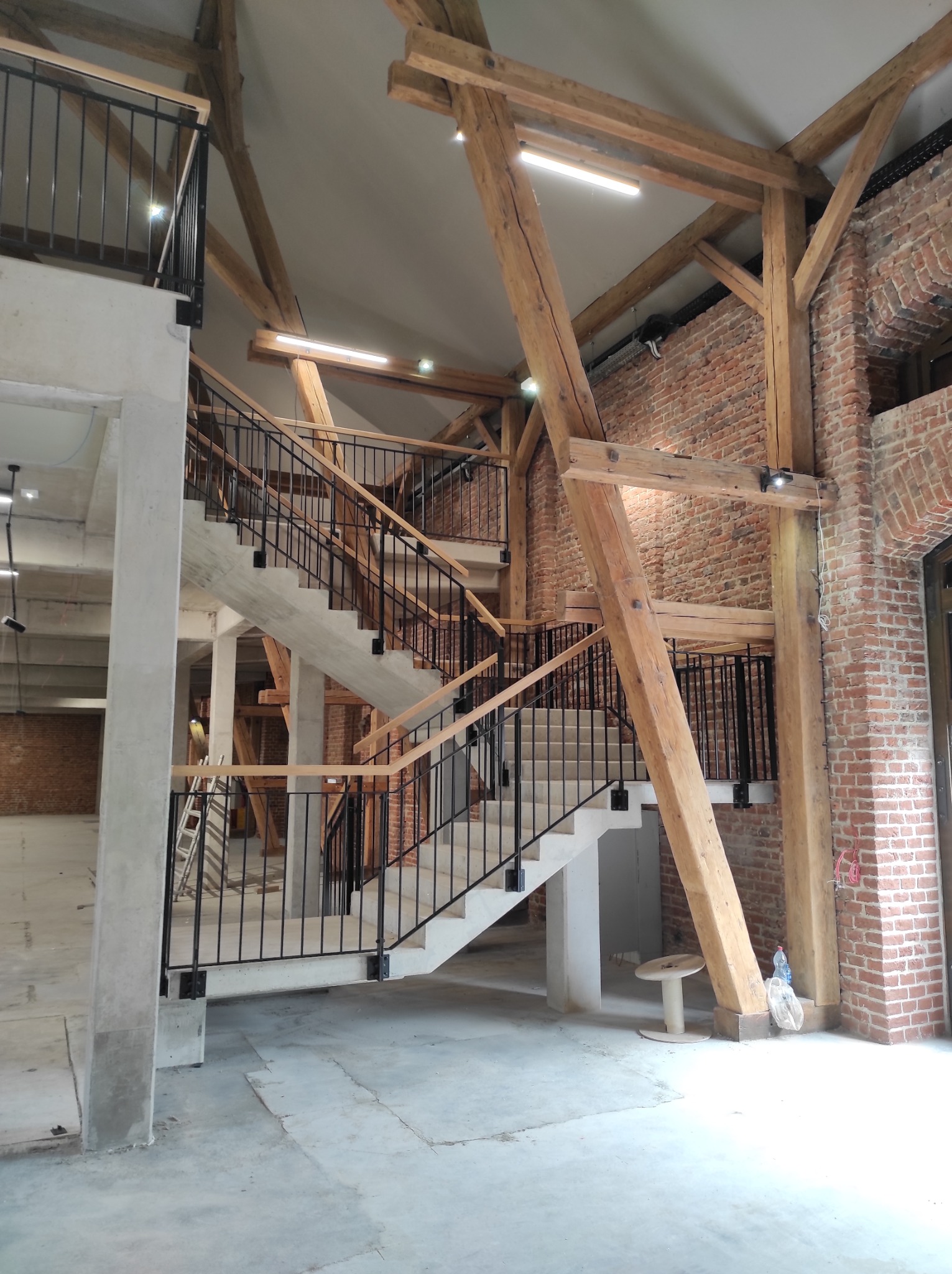 Wnętrze w trakcie wykończenia z betonowymi schodami, czarnymi balustradami i drewnianą konstrukcją stropu oraz fragmentem ściany z czerwonej cegły.