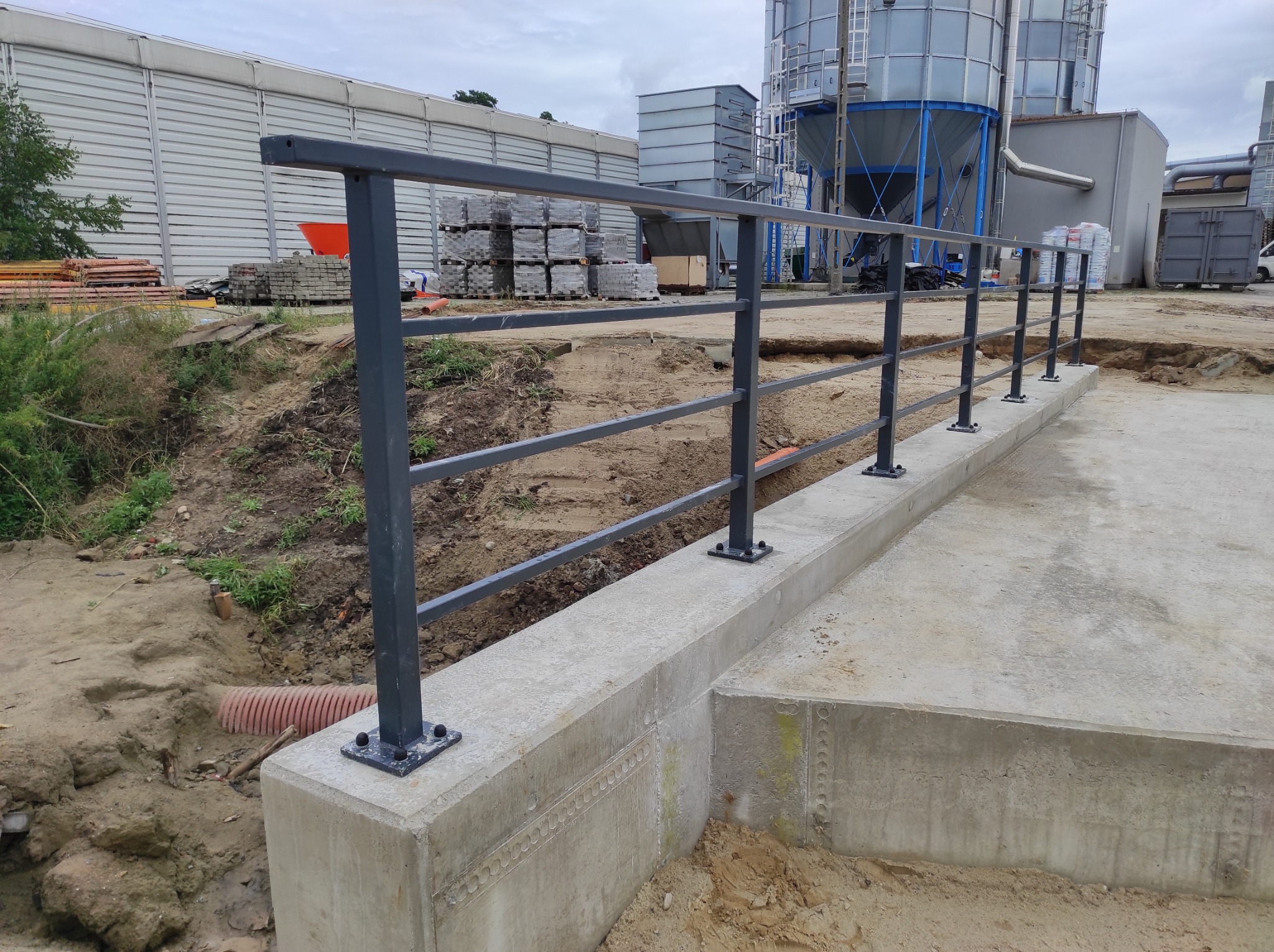 Szara metalowa barierka ochronna zamontowana na betonowym fundamencie, widoczna na tle placu budowy z zapleczem magazynowym i silosami.