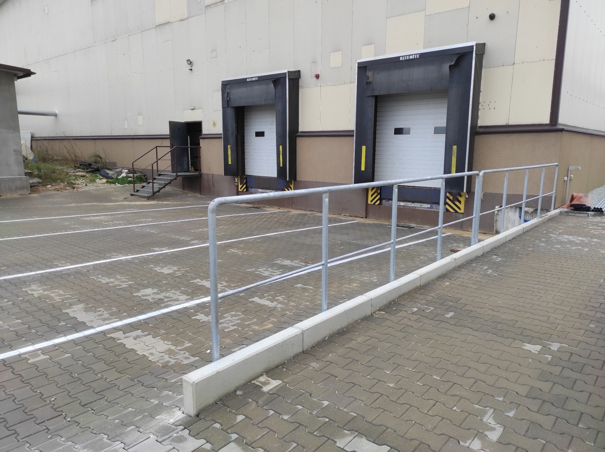Szara metalowa barierka ochronna przed budynkiem magazynowym z rampami załadunkowymi i betonową kostką brukową na parkingu.
