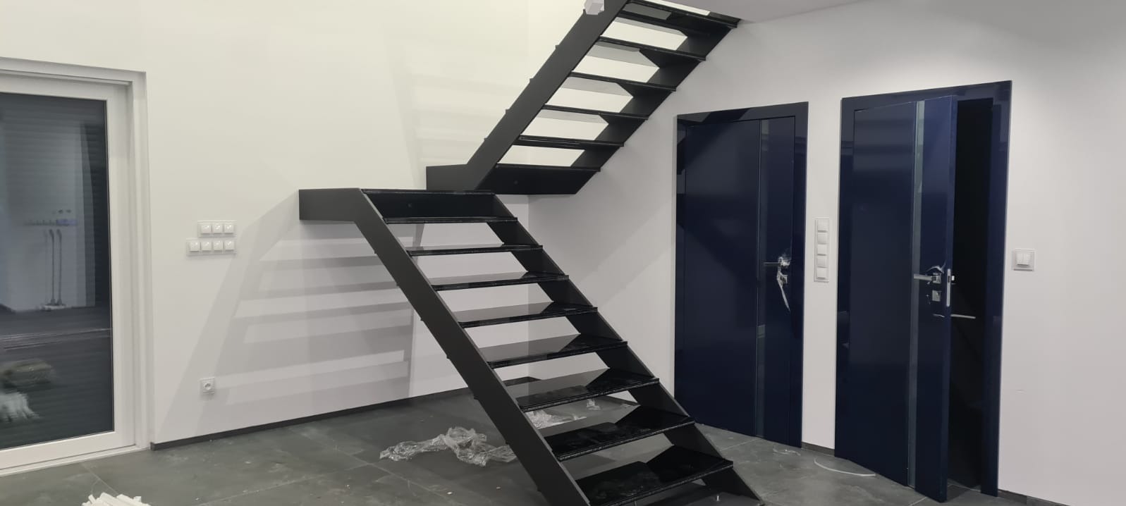 Nowoczesne, czarne, metalowe schody wewnętrzne z ażurowymi stopniami, zamontowane w minimalistycznym wnętrzu z białymi ścianami i ciemnymi drzwiami.