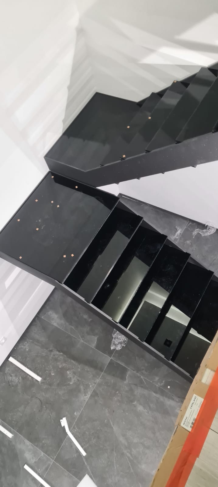 Nowoczesne, czarne schody wewnętrzne z metalową konstrukcją i szklanymi stopniami, widok z góry w trakcie montażu, widoczne ślady po naklejkach na szkle, szare płytki podłogowe, białe ściany.