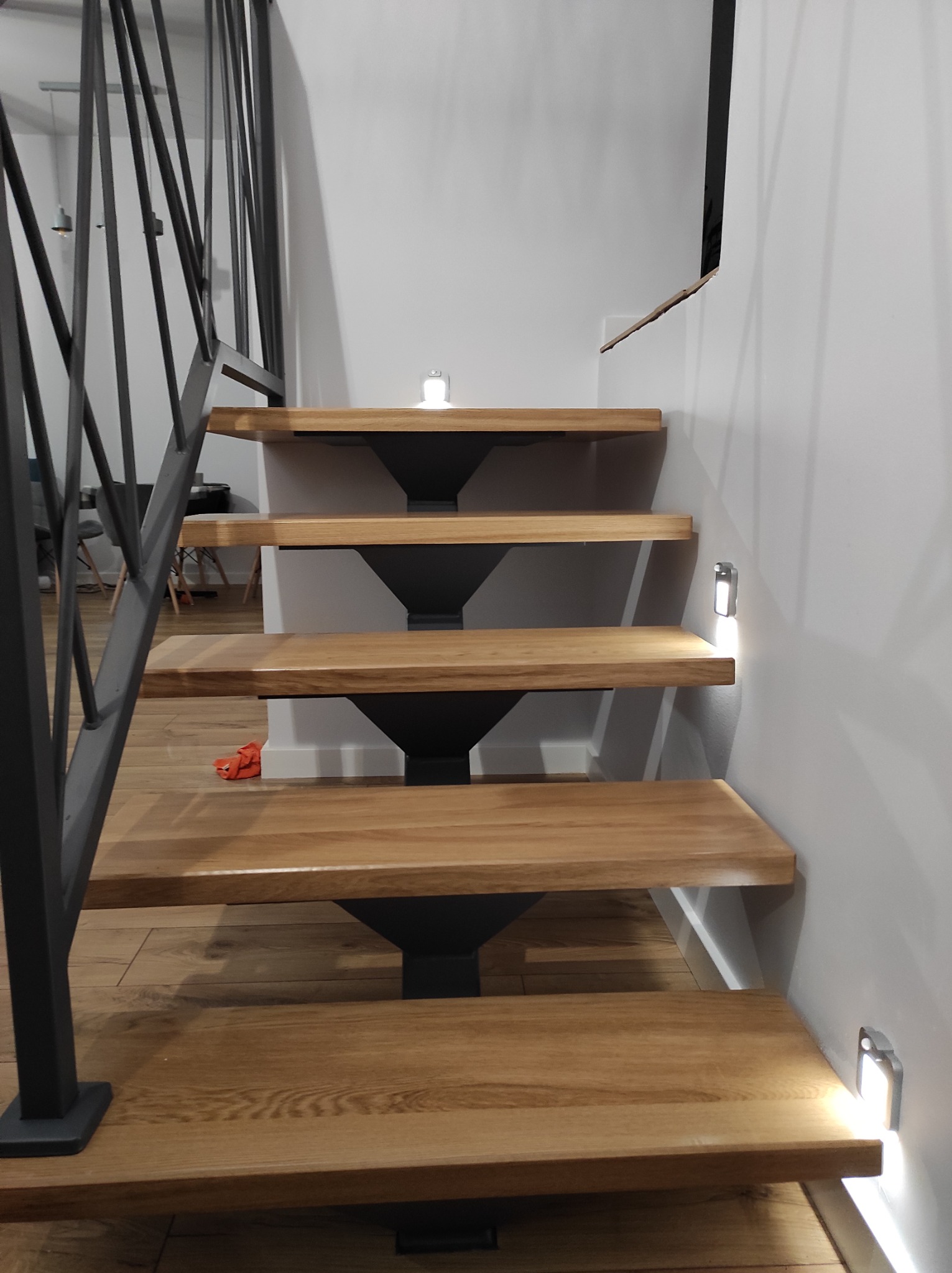 Schody drewniane z metalową balustradą o geometrycznym wzorze, oświetlenie punktowe na ścianie, widok od dołu.