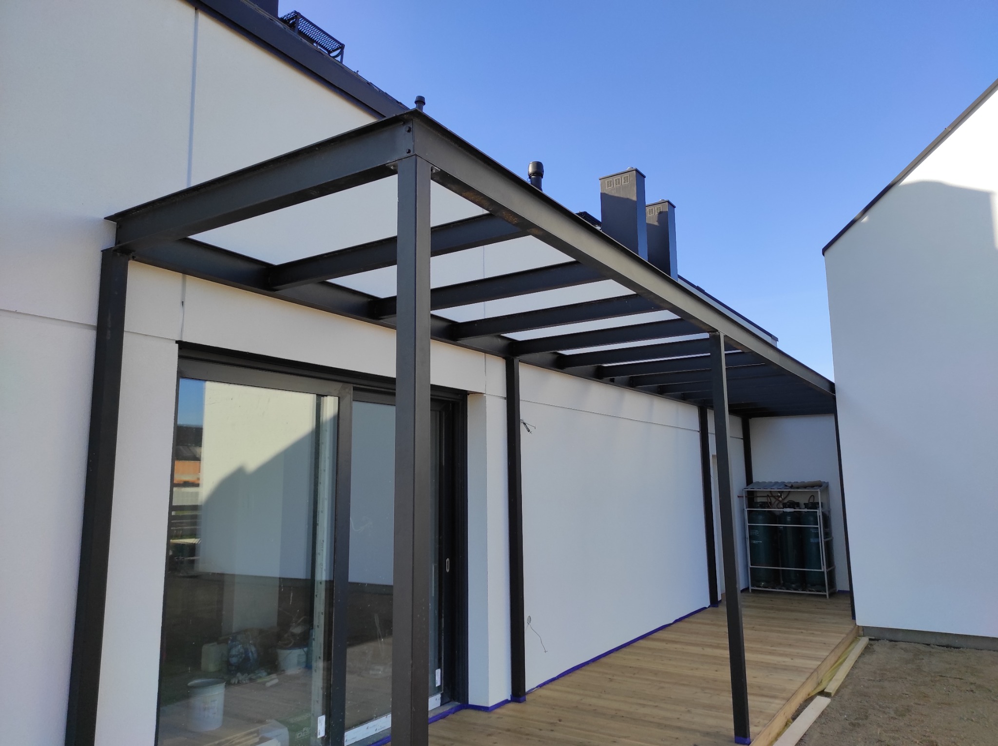Nowoczesna pergola z czarnej stali z przezroczystym zadaszeniem, przylegająca do białego budynku, widoczne butle z gazem w tle.
