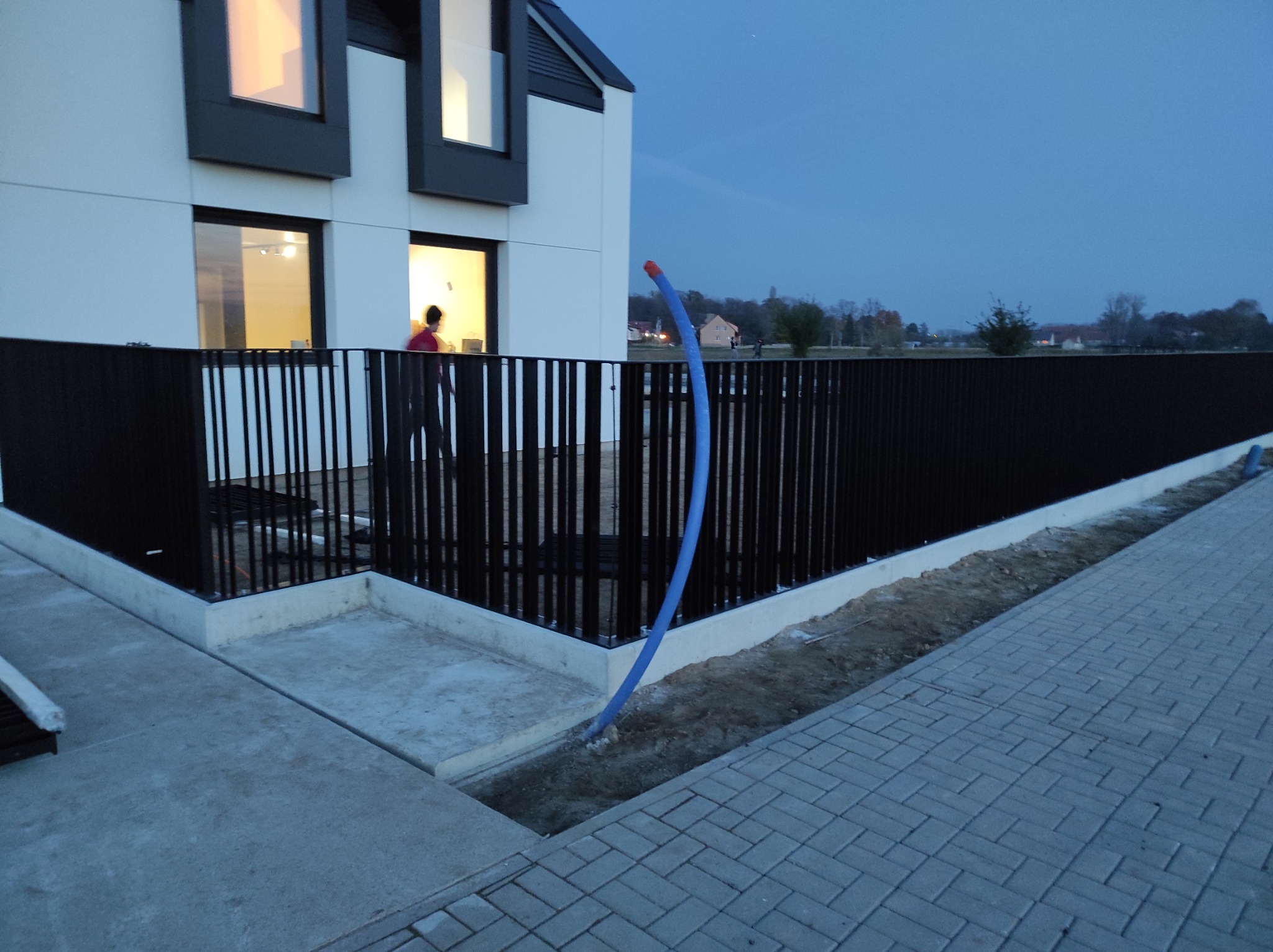 Nowoczesny dom z czarnym, metalowym ogrodzeniem sztachetowym, widoczne betonowe obrzeże i niebieski przewód wystający z ziemi, kostka brukowa w tle, wieczorne oświetlenie