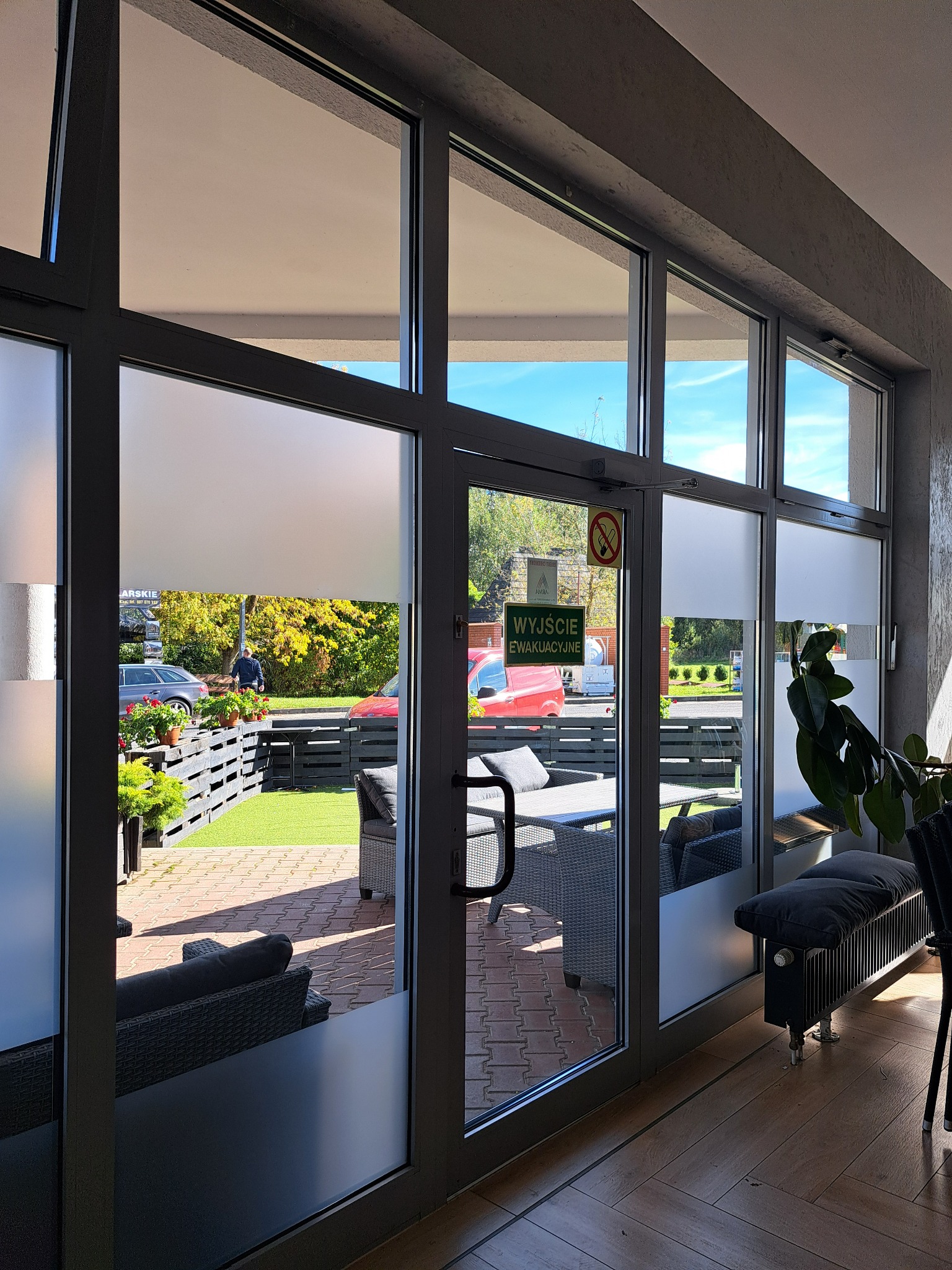 Widok przez przeszklone drzwi na patio z meblami ogrodowymi, trawnikiem i roślinami w donicach, częściowo zasłonięty matową folią na szybach.