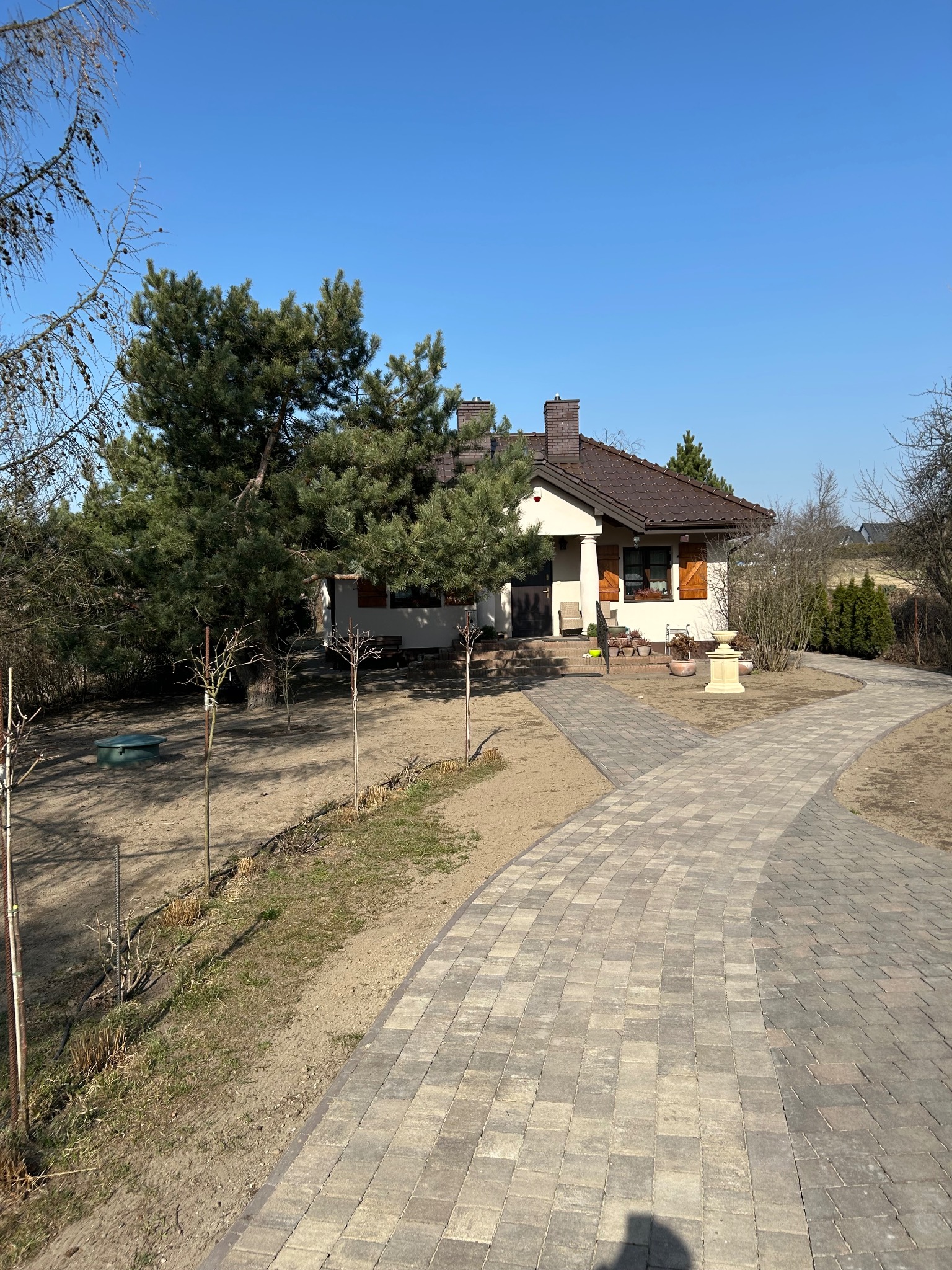 Ułożona kostka brukowa prowadząca do domu z ogrodem, widoczne drzewa iglaste i elementy dekoracyjne ogrodu, błękitne niebo w tle.