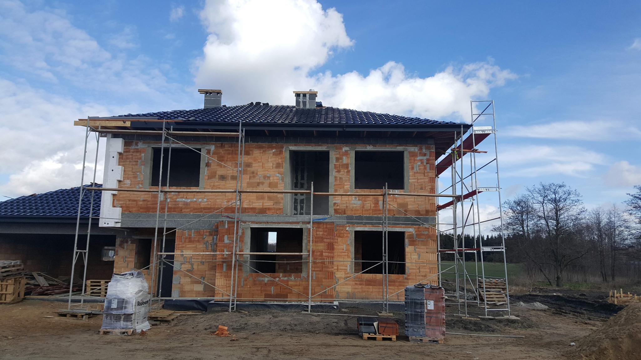 Dwukondygnacyjny dom w trakcie budowy z cegły ceramicznej, z widocznymi rusztowaniami, niebieską dachówką i przygotowanym materiałem izolacyjnym, na tle błękitnego nieba z chmurami.