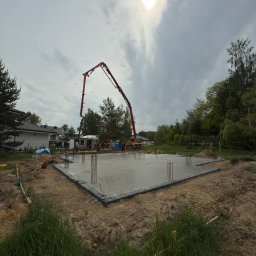 Projekty i Nadzory Budowlane Projektivo - Wylewka fundamentów domu jednorodzinnego z użyciem pompy do betonu. Widoczne wystające pręty zbrojeniowe i pracownicy w tle. Pochmurny dzień.