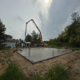 Wylewka fundamentów domu jednorodzinnego z użyciem pompy do betonu. Widoczne wystające pręty zbrojeniowe i pracownicy w tle. Pochmurny dzień.