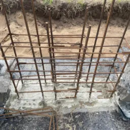 Zbrojenie fundamentu pod budowę, widoczne pręty stalowe połączone w siatkę, zalane betonem. W tle ziemia i trawa. Detal z bliska ukazuje etap prac budowlanych.