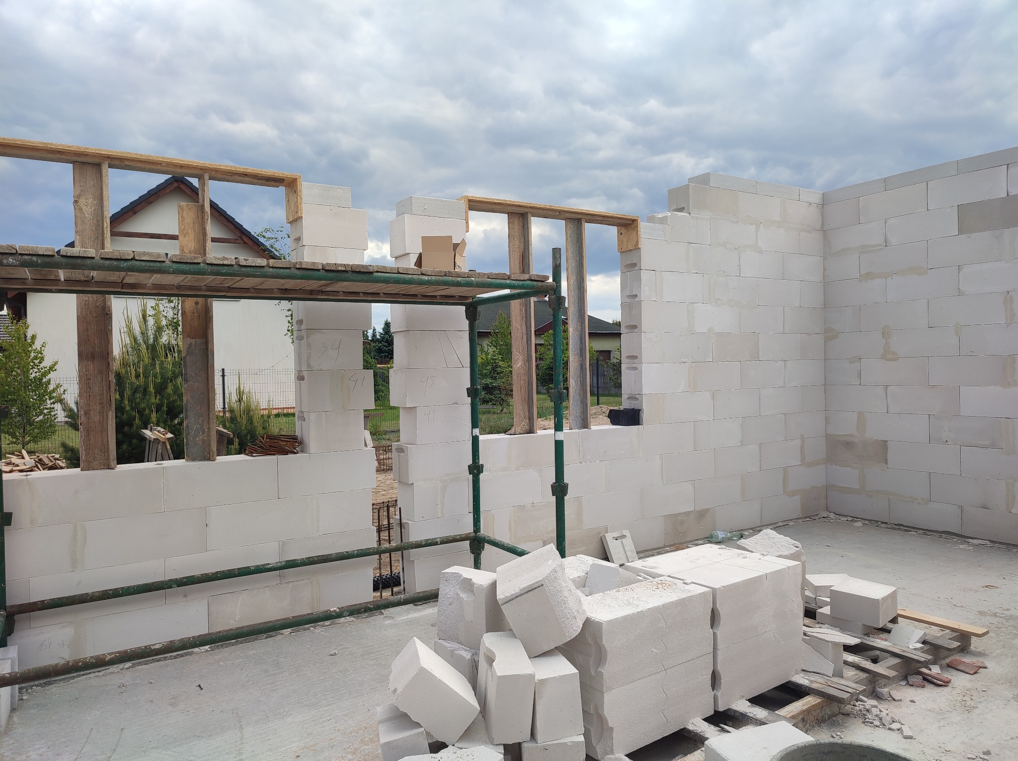 Wznoszenie ścian domu z białych bloczków, widoczne rusztowanie i otwory okienne. Część bloczków leży na podłożu. Tło: pochmurne niebo i fragmenty zabudowy.