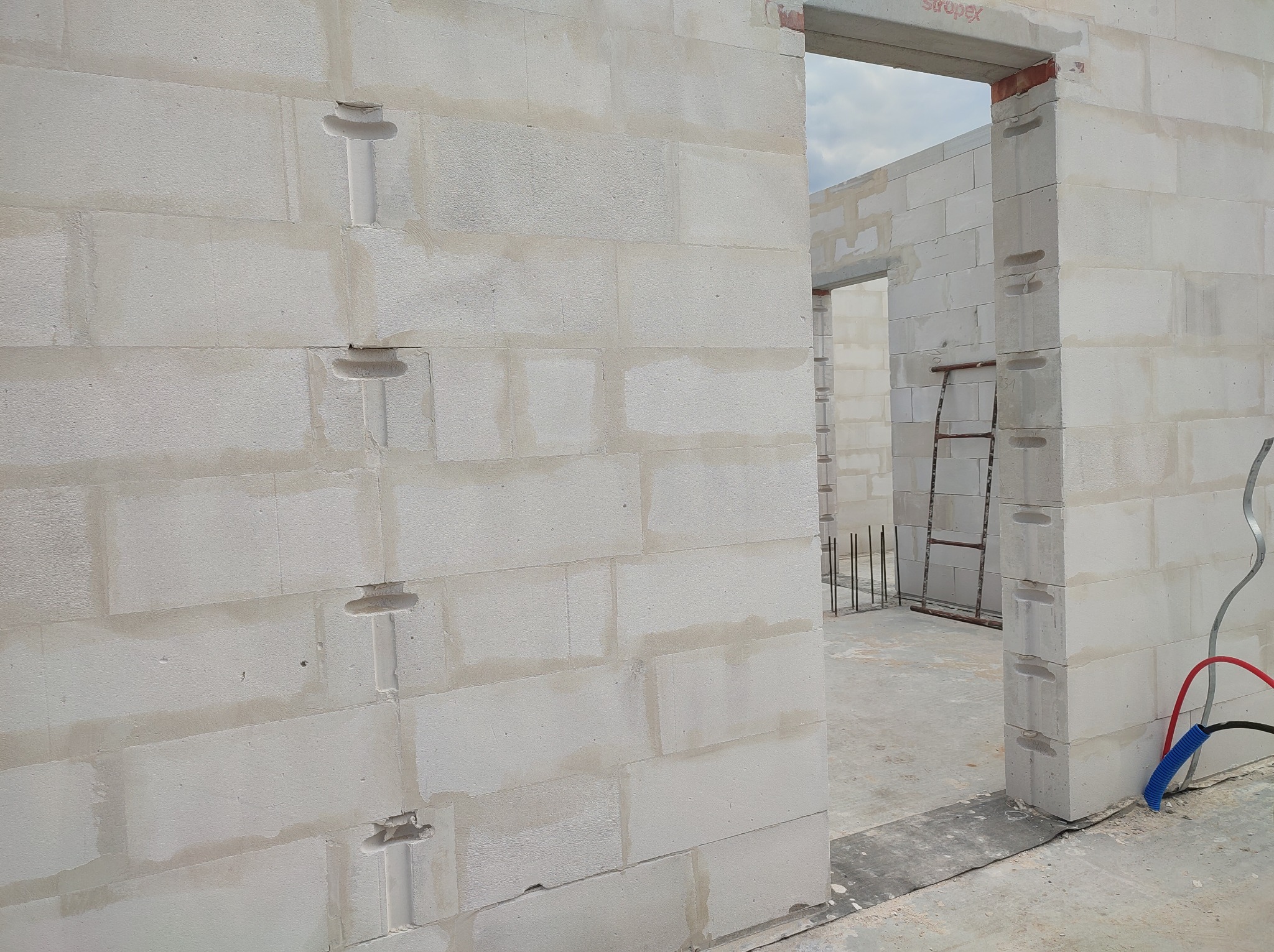 Murowane ściany z betonu komórkowego w surowym stanie, widoczne otwory instalacyjne i przewody elektryczne. W tle drabina i pręty zbrojeniowe.