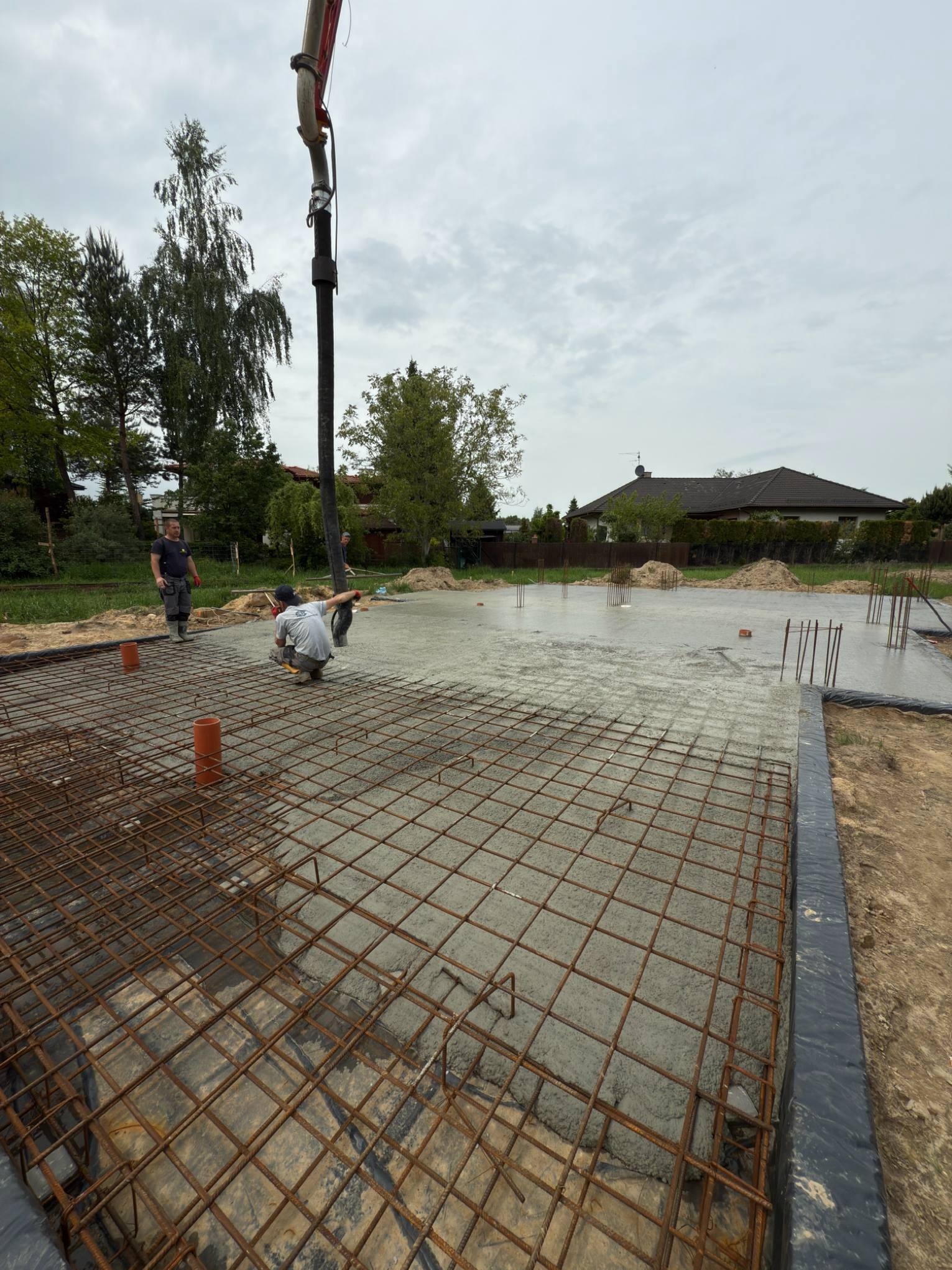 Wylewanie betonu na zbrojenie fundamentu domu jednorodzinnego. Widoczni pracownicy budowlani i pompa do betonu. Tło stanowią drzewa i dom.