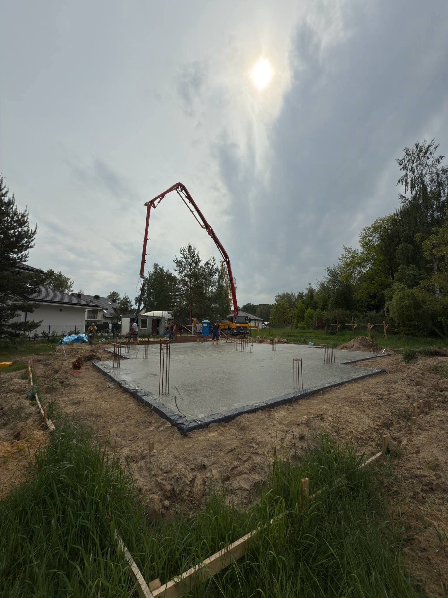 Wylewka fundamentów domu jednorodzinnego z użyciem pompy do betonu. Widoczne wystające pręty zbrojeniowe i pracownicy w tle. Pochmurny dzień.