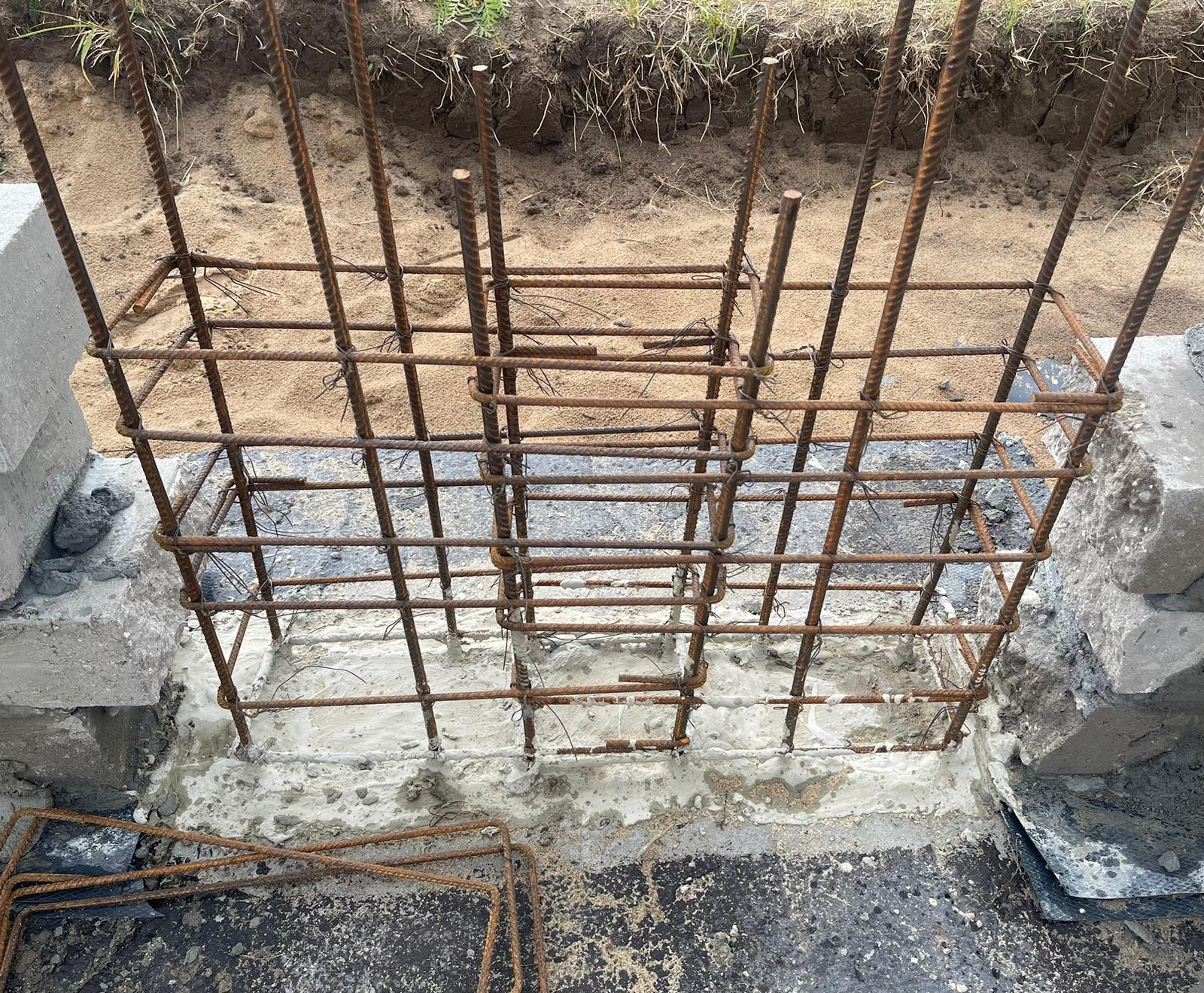 Zbrojenie fundamentu pod budowę, widoczne pręty stalowe połączone w siatkę, zalane betonem. W tle ziemia i trawa. Detal z bliska ukazuje etap prac budowlanych.