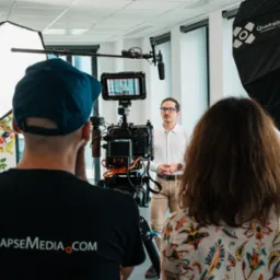 Ekipa filmowa podczas kręcenia ujęcia z mężczyzną w białej koszuli, widoczny monitor kamery, softbox Quadralite i logo TimeLapseMedia.com na koszulce operatora.