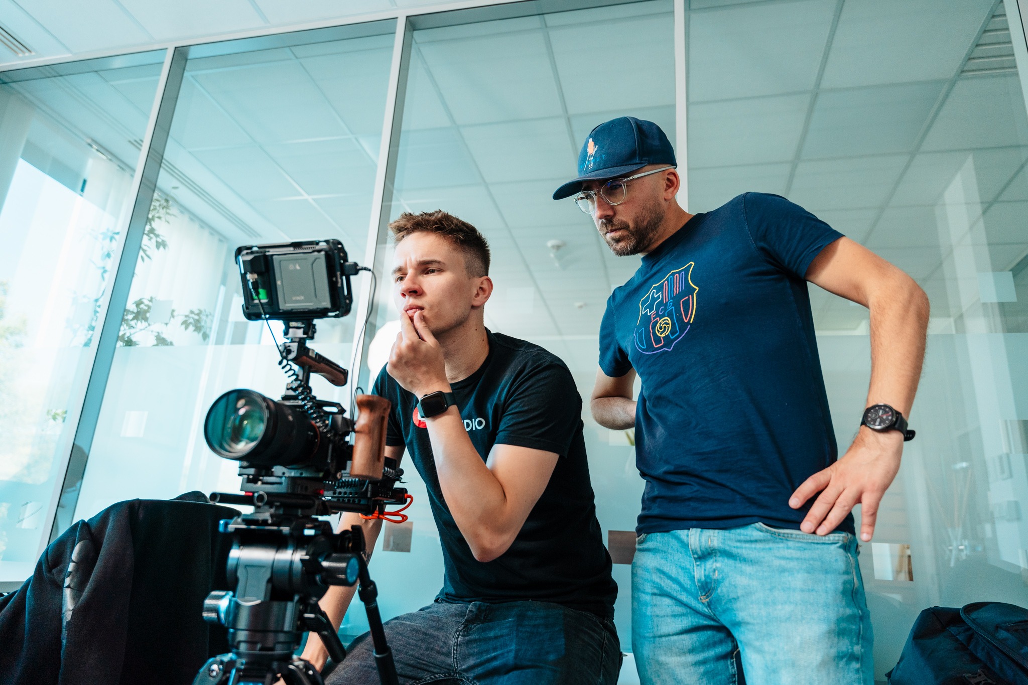 Dwaj mężczyźni w studio filmowym, jeden siedzi i patrzy na ekran kamery, drugi stoi obok z ręką na biodrze, obaj ubrani w codzienne ubrania, na pierwszym planie profesjonalna kamera na statywie.