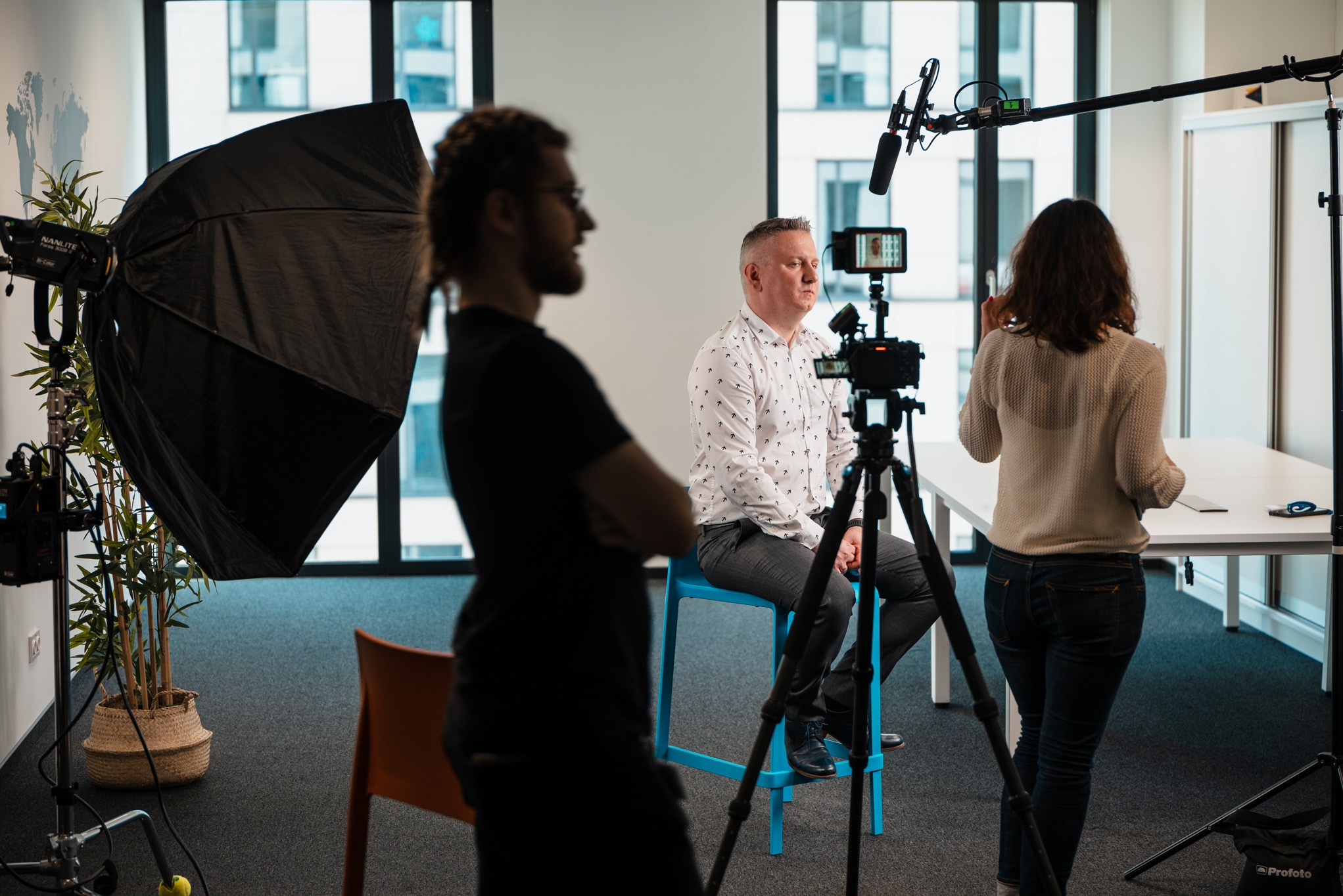Plan filmowy w biurze: mężczyzna siedzi na niebieskim krześle, oświetlony lampą, nagrywany przez kamerę z mikrofonem, z operatorem i asystentem w tle.