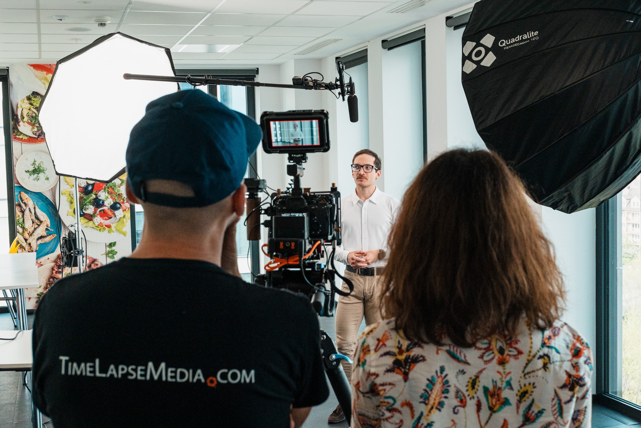 Ekipa filmowa podczas kręcenia ujęcia z mężczyzną w białej koszuli, widoczny monitor kamery, softbox Quadralite i logo TimeLapseMedia.com na koszulce operatora.