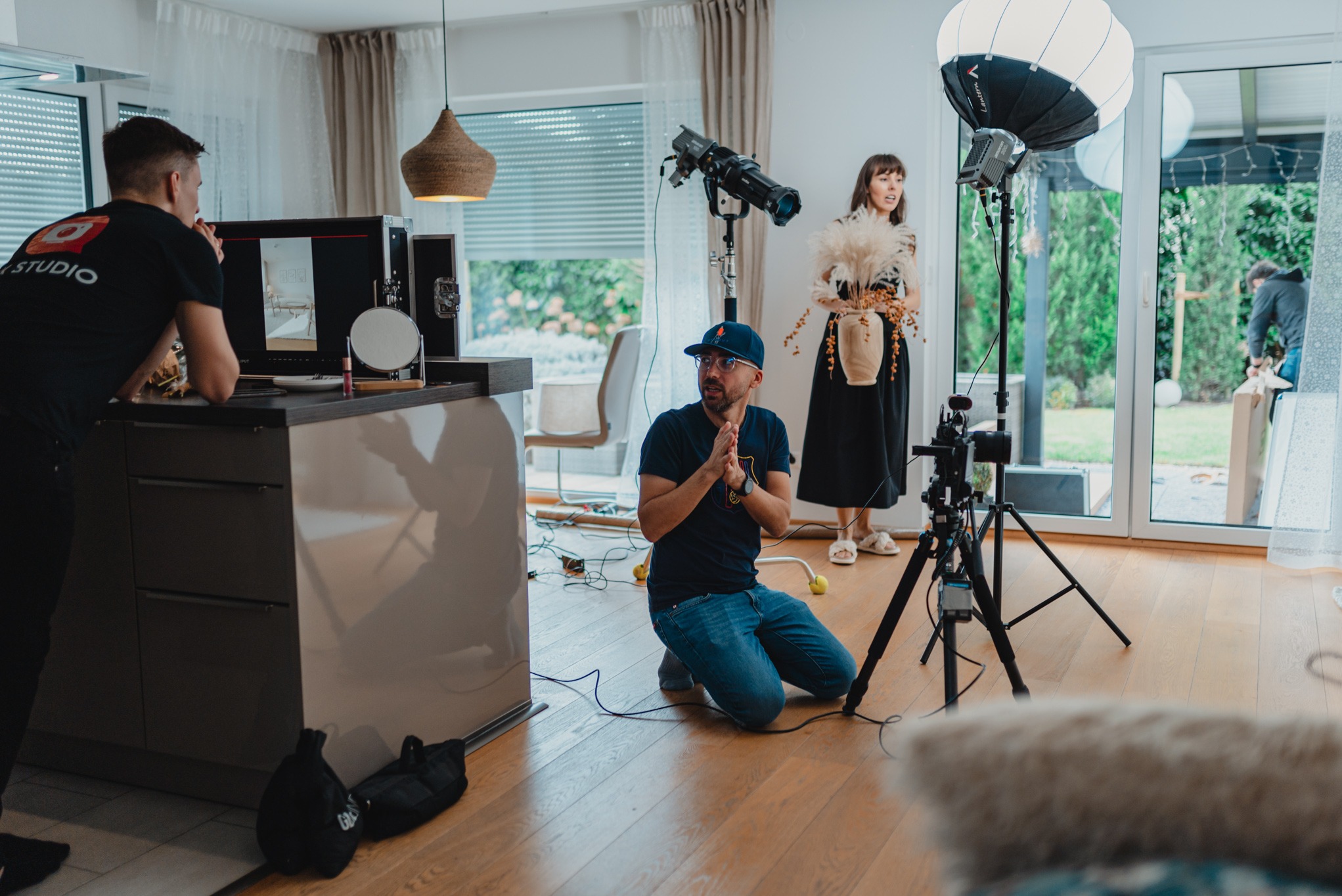 Ekipa filmowa podczas sesji w jasnym wnętrzu: modelka z wazonem, operator na kolanach, asystent przy monitorze, oświetlenie studyjne.