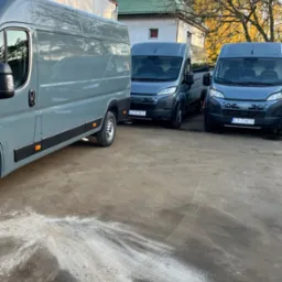 Trzy szare busy Fiat Ducato zaparkowane na placu, z widocznymi tablicami rejestracyjnymi, w tle budynek z zielonym dachem i drzewa z jesiennymi liśćmi.