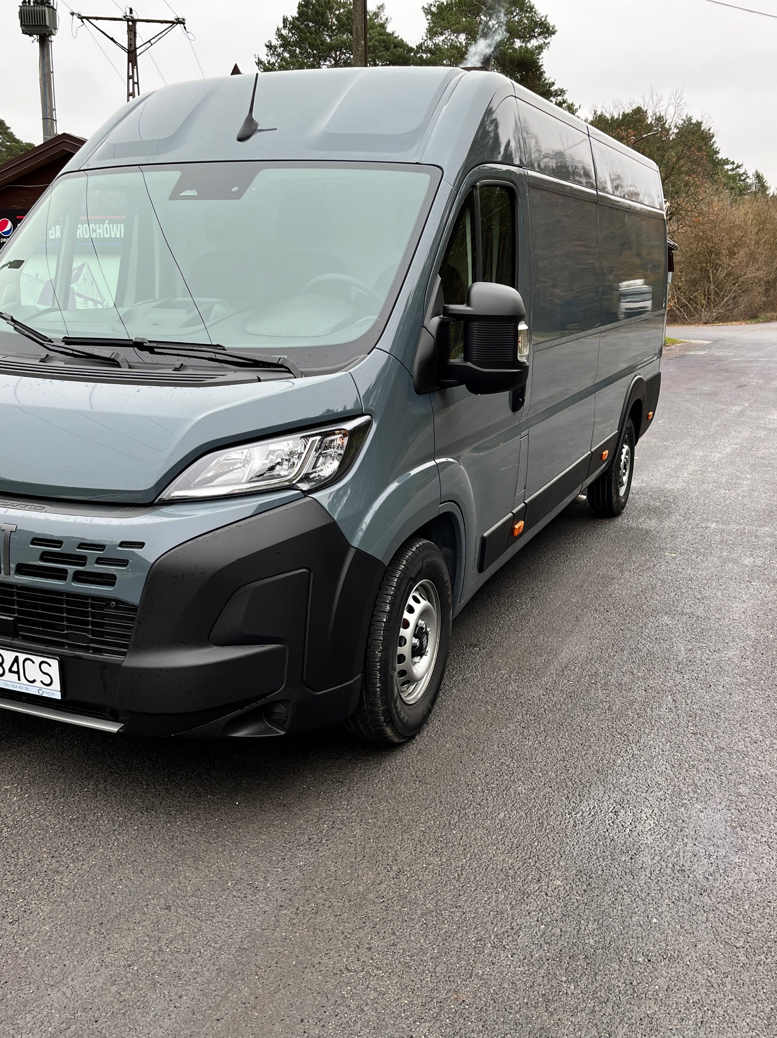 Szary, nowoczesny bus dostawczy zaparkowany na mokrym asfalcie, widok z przodu na reflektor i tablicę rejestracyjną, w tle drzewa i budynek z kominem, z którego unosi się dym.