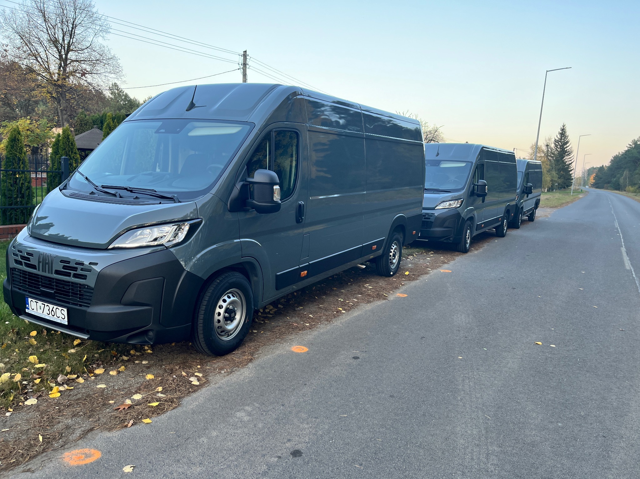 Szereg identycznych, szarych busów Fiat Ducato zaparkowanych wzdłuż drogi, z widoczną tablicą rejestracyjną pierwszego pojazdu i pomarańczowymi oznaczeniami na asfalcie.
