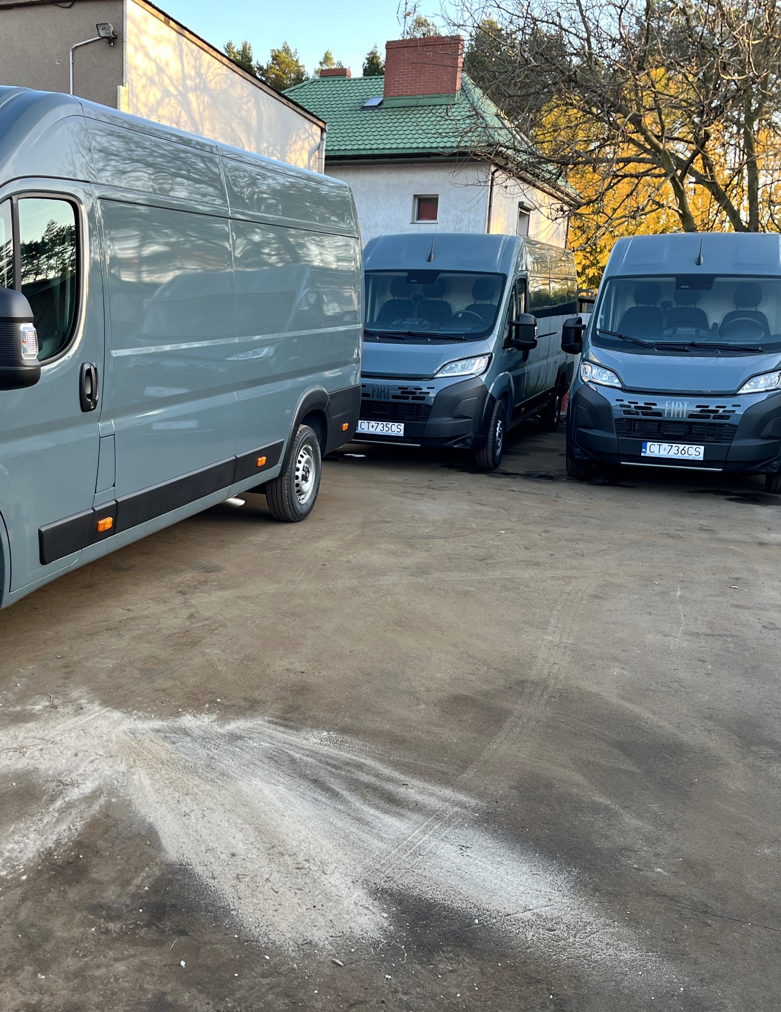 Trzy szare busy Fiat Ducato zaparkowane na placu, z widocznymi tablicami rejestracyjnymi, w tle budynek z zielonym dachem i drzewa z jesiennymi liśćmi.