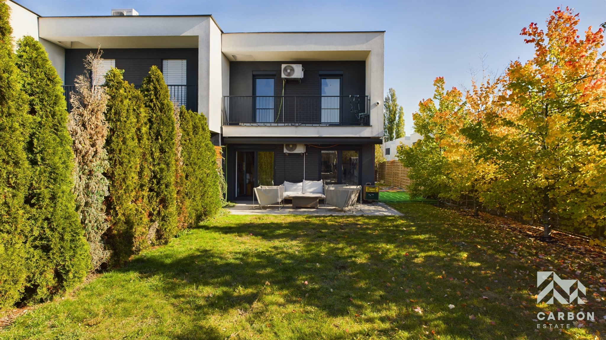 Nowoczesny, dwukondygnacyjny dom z ogrodem w Chorzowie, z balkonem i tarasem. Widoczne krzewy i drzewa w jesiennych barwach. Minimalistyczny design.