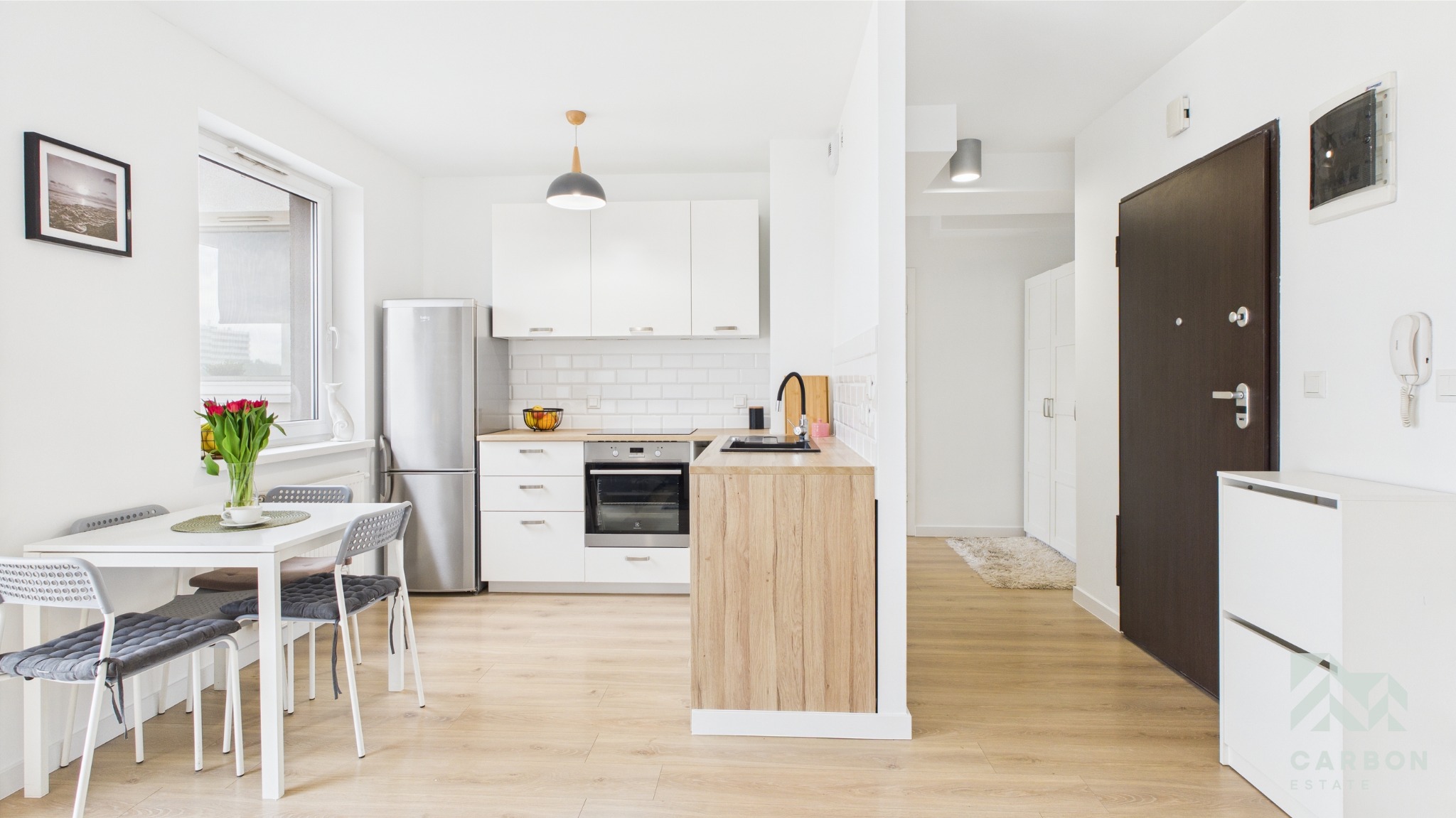 Jasne wnętrze mieszkania: kuchnia z jadalnią i przedpokój. Białe meble, drewniane blaty, szare krzesła. Widok na drzwi wejściowe i szafę. Nowoczesny design.