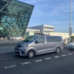 Bus4_you - Srebrny minivan Toyota Proace Verso zaparkowany przed budynkiem lotniska Kraków Airport, widoczne tablice rejestracyjne, nowoczesna architektura w tle.