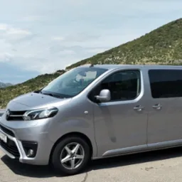 Srebrny van Toyota Proace Verso z przyciemnianymi szybami na tle górskiego krajobrazu. Auto zaparkowane na poboczu, widok z boku, lekko z góry. Jasne, naturalne światło.