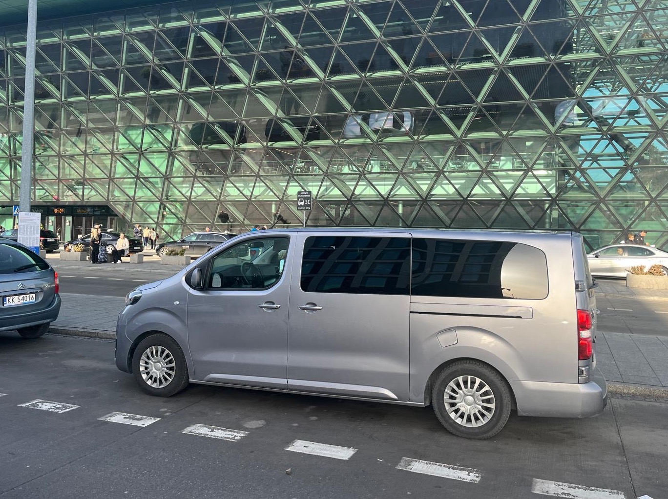 Szary van osobowy zaparkowany przed nowoczesnym budynkiem lotniska z geometryczną fasadą, widoczne inne samochody i osoby w tle.