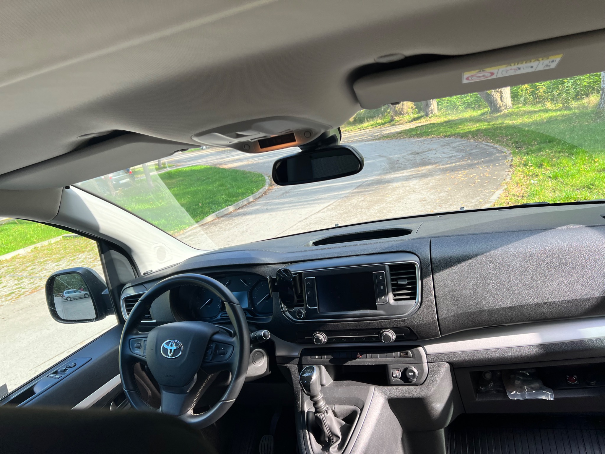 Wnętrze samochodu dostawczego marki Toyota, widok z perspektywy kierowcy, z widoczną deską rozdzielczą, kierownicą z logo, lusterkiem wstecznym i fragmentem drogi z zielenią za szybą.