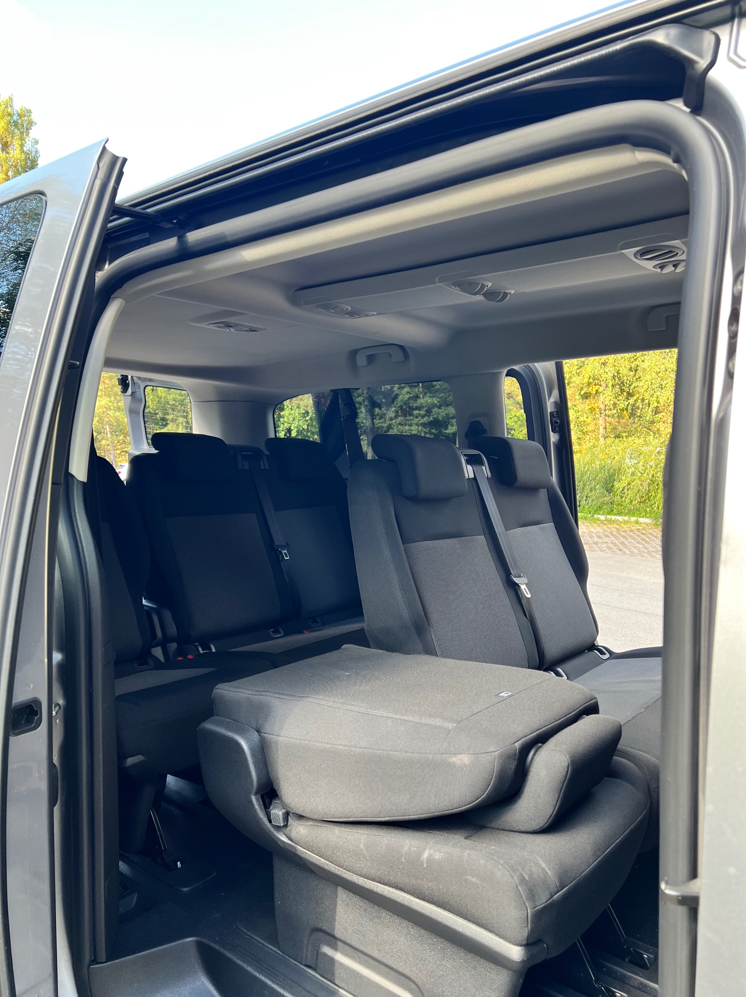 Wnętrze busa z rozłożonymi siedzeniami, widok z boku przez otwarte drzwi, szara tapicerka.