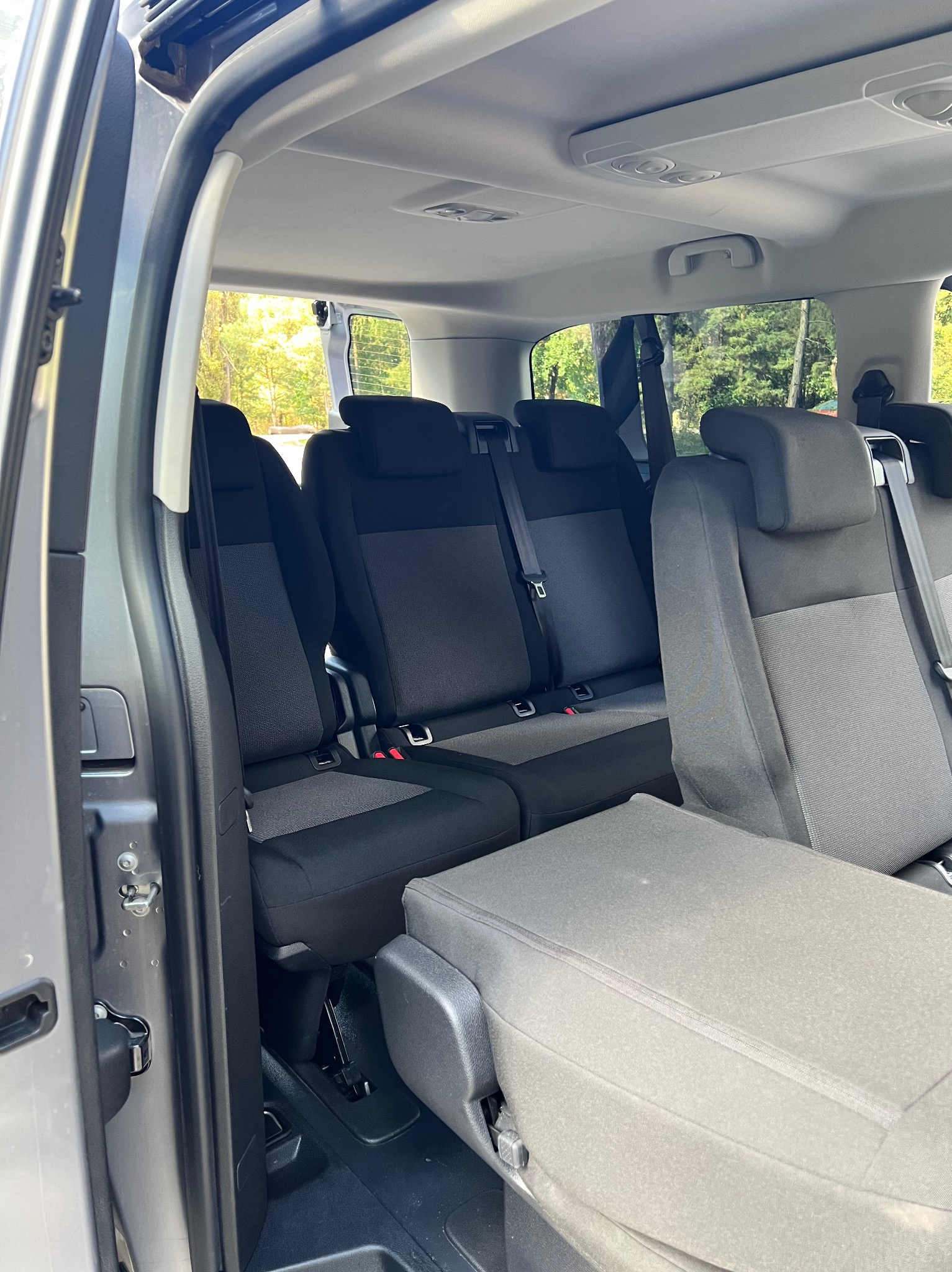 Wnętrze szarego busa z trzema rzędami siedzeń, widok z otwartych drzwi bocznych, złożone siedzenie w pierwszym planie.