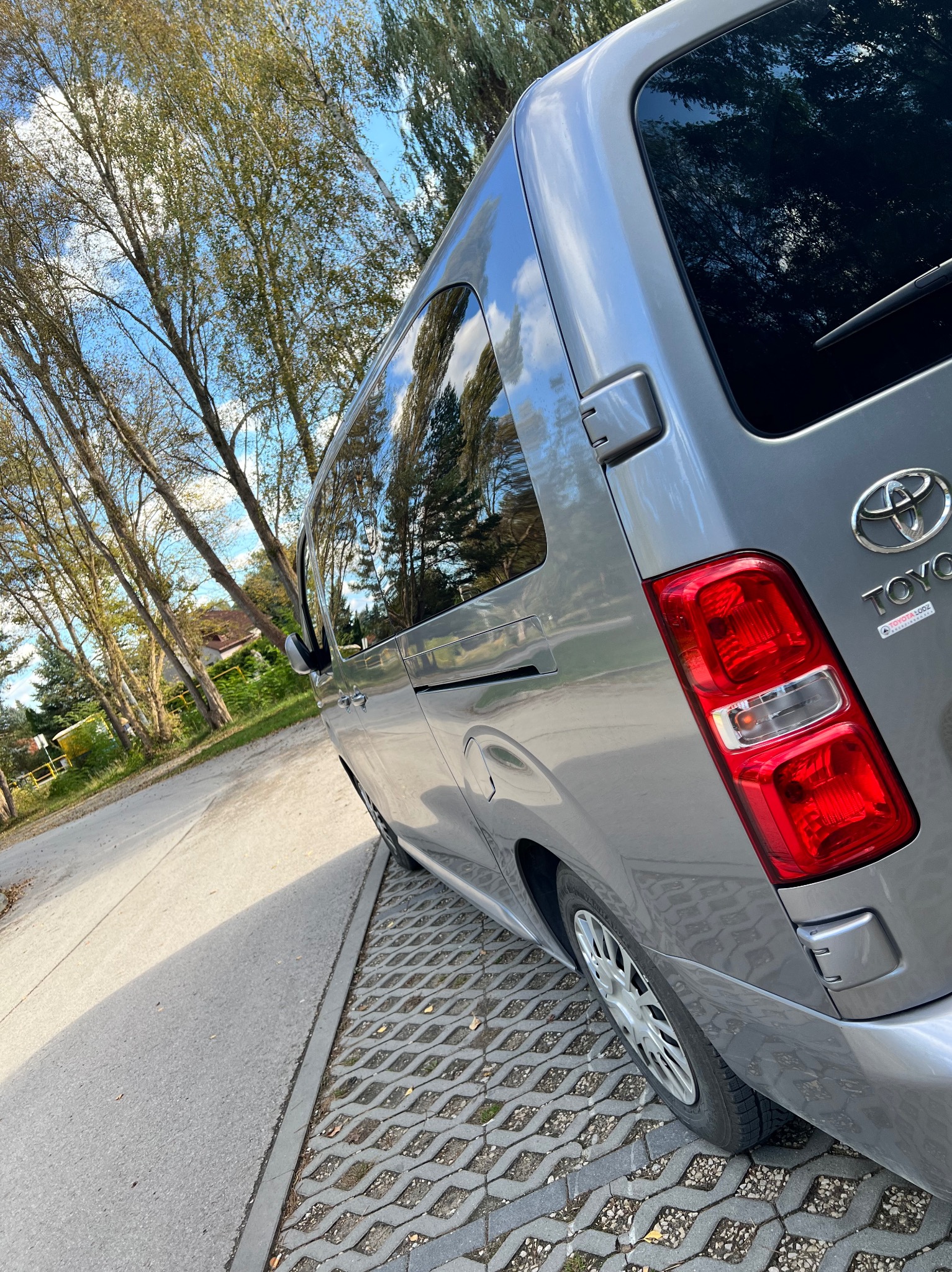 Szary van Toyota z widocznym logo producenta, zaparkowany na betonowej kostce ażurowej, z drzewami odbijającymi się w szybach.