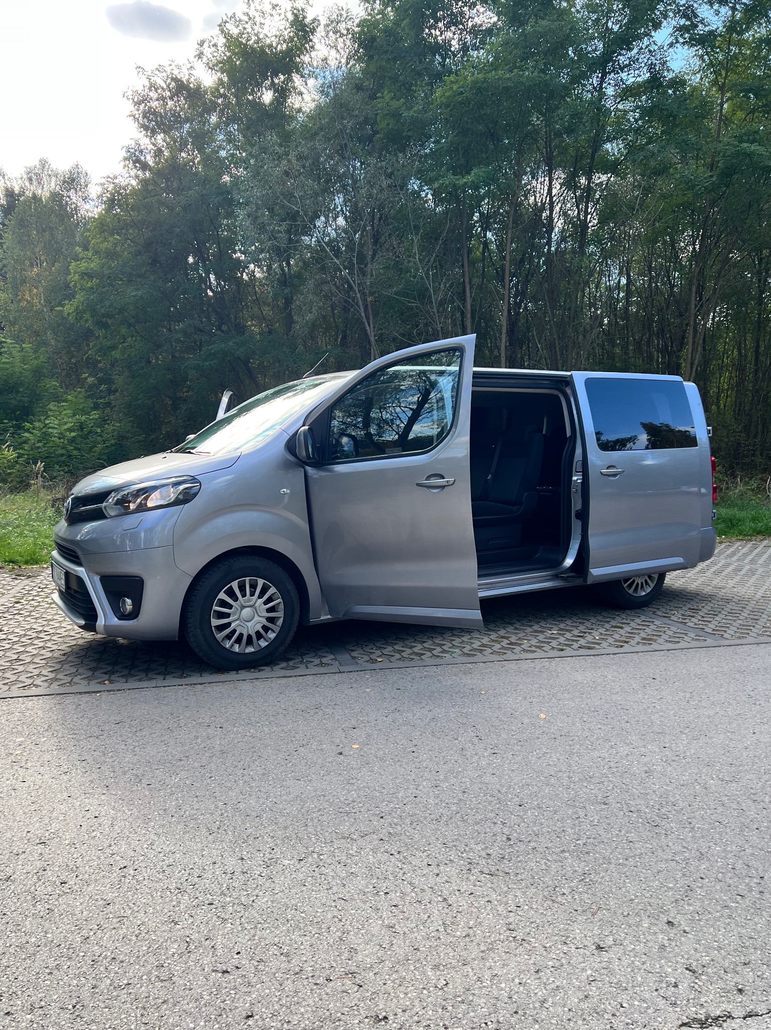 Srebrny van z otwartymi drzwiami przesuwnymi, zaparkowany na parkingu z kostki, z zielonymi drzewami w tle.