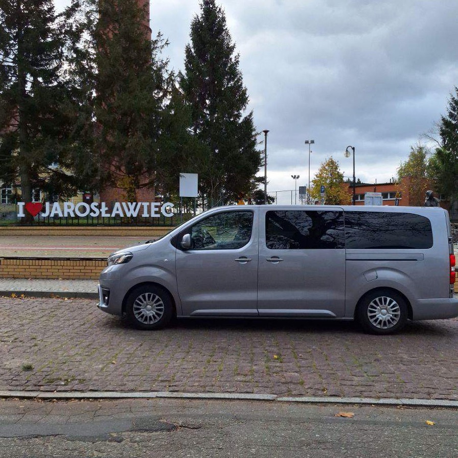 Srebrny van zaparkowany na brukowanej ulicy z napisem 'I love Jarosławiec' w tle, pochmurne niebo.