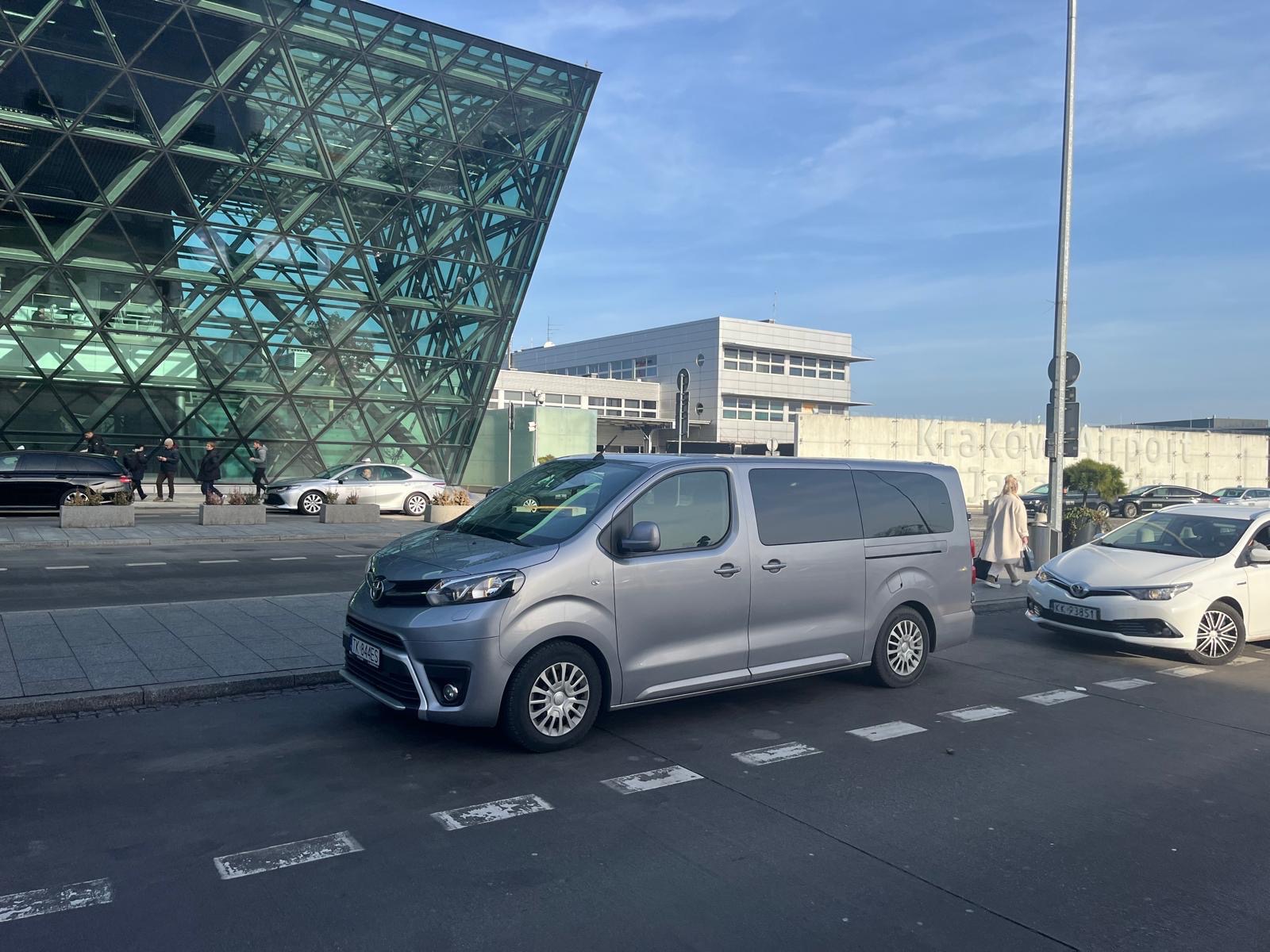 Srebrny minivan Toyota Proace Verso zaparkowany przed budynkiem lotniska Kraków Airport, widoczne tablice rejestracyjne, nowoczesna architektura w tle.